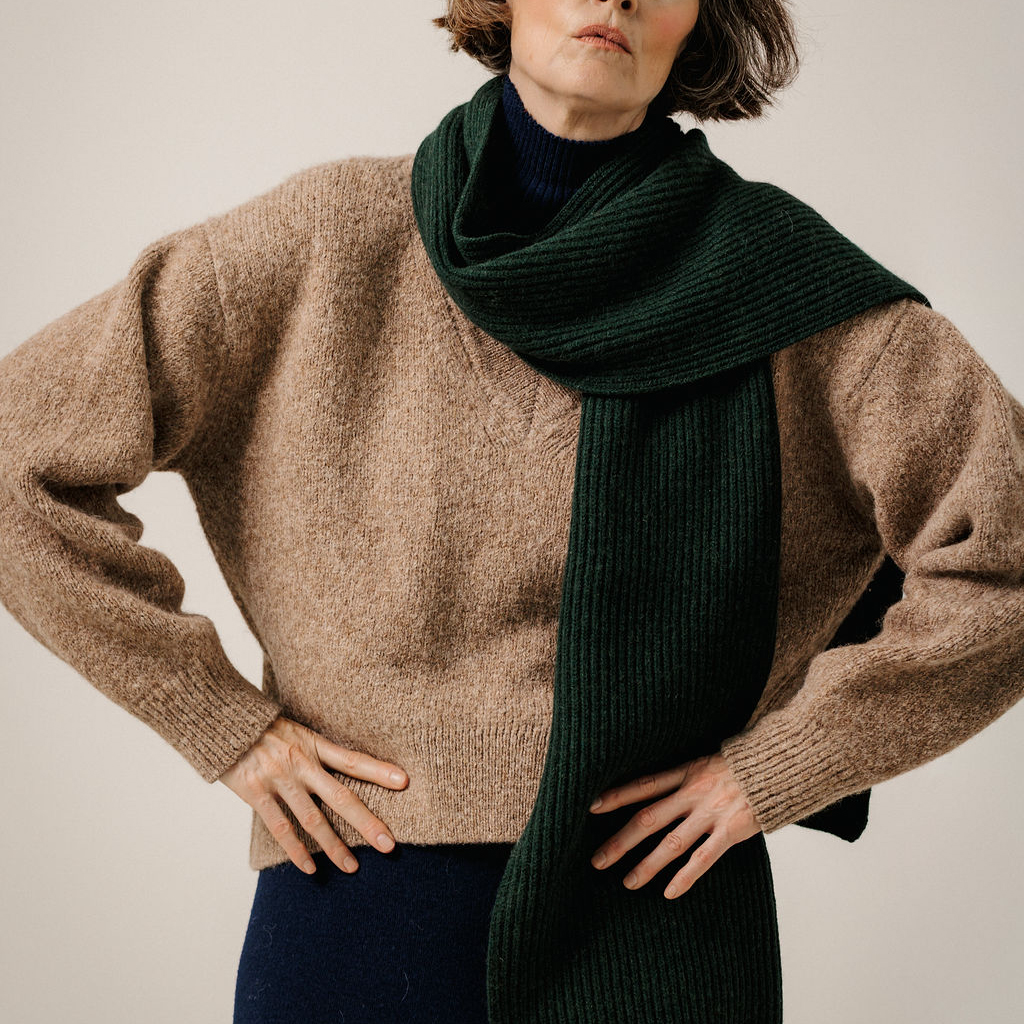 A model wearing a chunky knit beige sweater and a dark forest green ribbed scarf, photographed against a neutral light beige background.