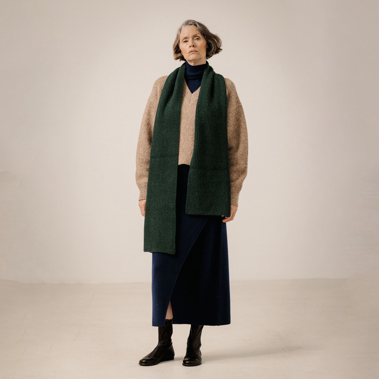 A woman stands in a studio wearing a long dark green ribbed knit scarf over a beige sweater, navy turtleneck, navy skirt, and black ankle boots.