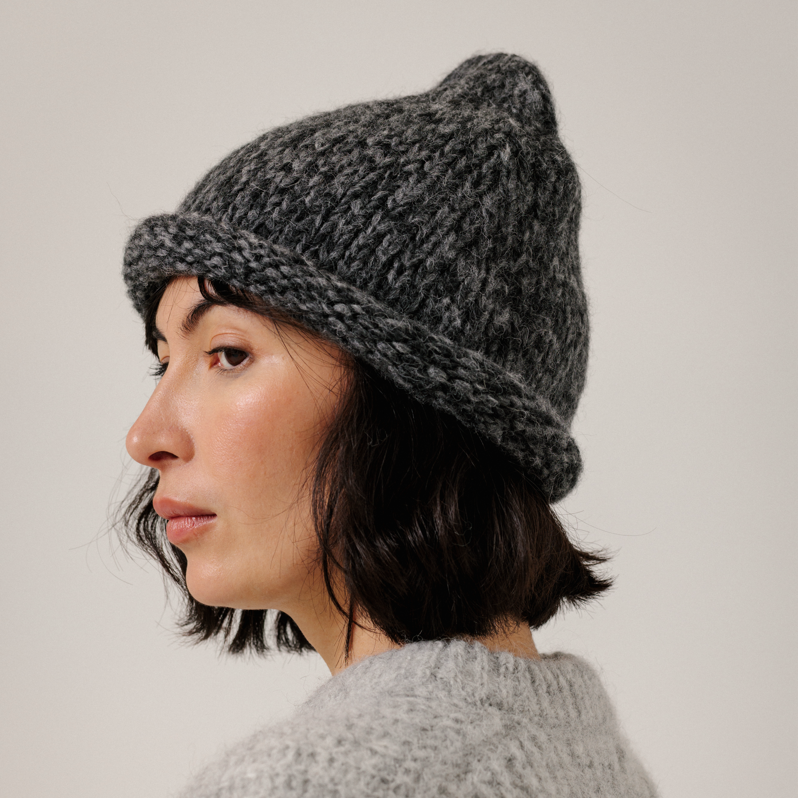 A woman shown in profile wearing a dark gray chunky knit beanie hat and a light gray knit sweater, photographed against a plain light gray background.