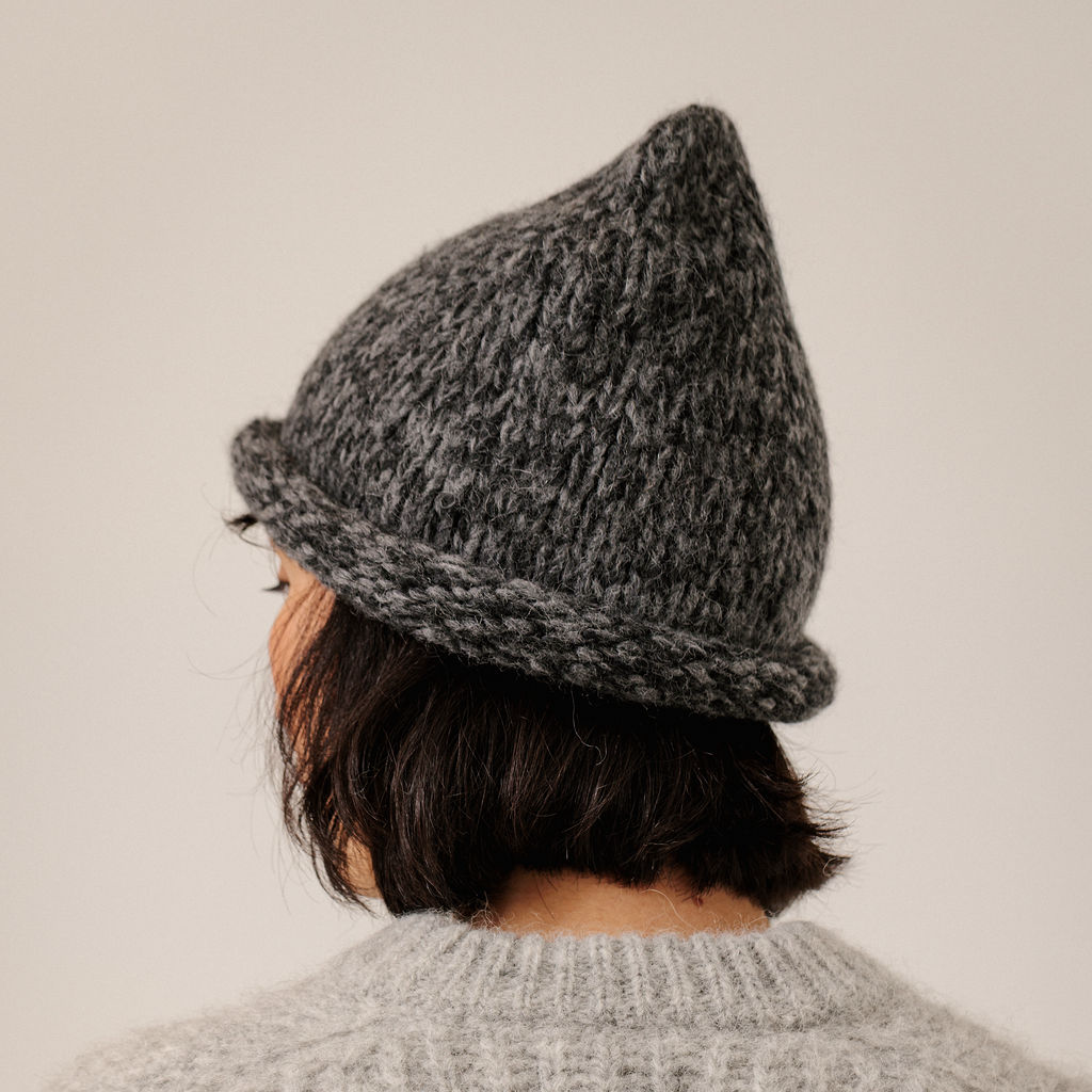 A person shown from the back wearing a dark gray marled knit beanie with a rolled brim and a chunky knit light gray sweater, against a plain light beige background.