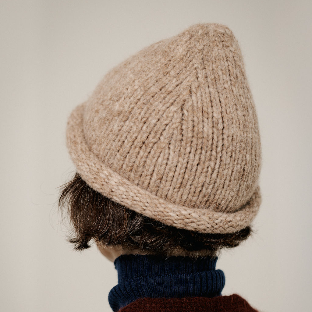 A close-up back view of a person wearing a chunky knit beanie in a warm beige/tan color, paired with a dark navy turtleneck sweater against a plain light background.