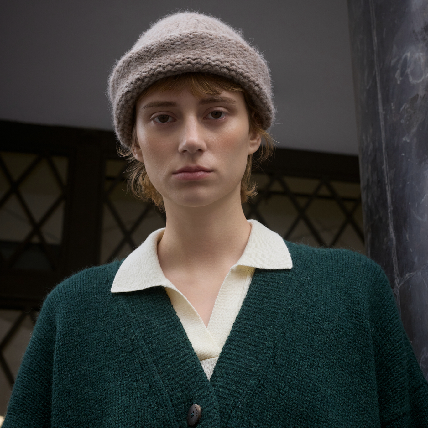 A person wearing a chunky knit beige beanie, a dark green cardigan over a cream collared shirt, standing indoors near a dark pillar.