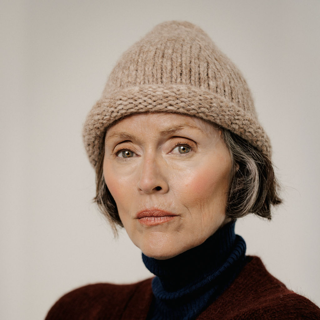 A close-up portrait of a woman wearing a beige chunky knit beanie hat, a dark navy turtleneck, and a maroon sweater against a plain light gray background.