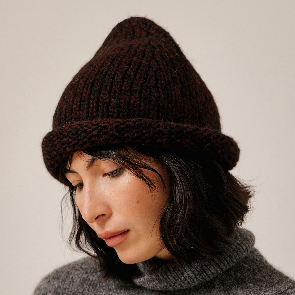 A close-up of a woman wearing a dark maroon chunky knit beanie hat and a gray turtleneck sweater, set against a plain light beige background.