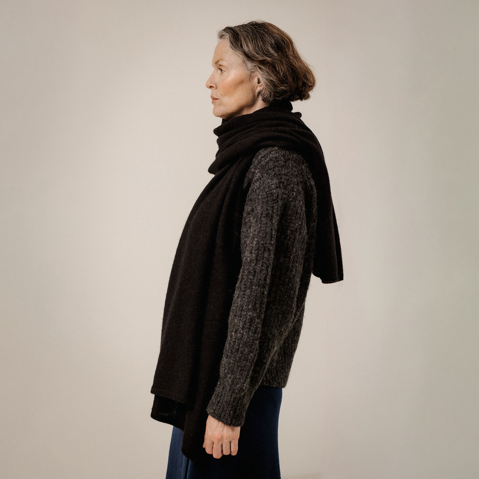 A woman in profile wearing a dark brown knit sweater and a black draped shawl, photographed against a plain light beige studio background.