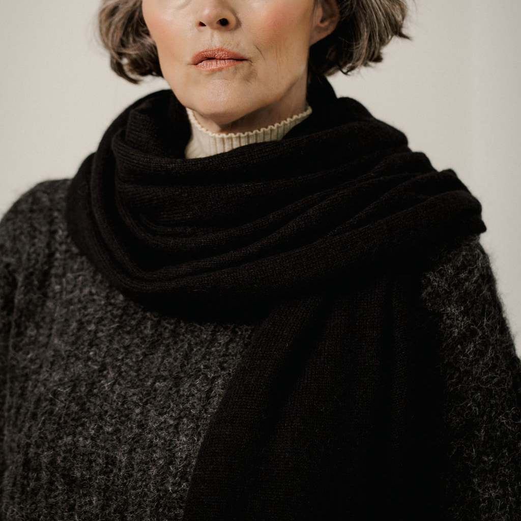 A close-up of a woman wearing a thick black knit scarf wrapped around her neck, layered over a dark gray textured sweater and a cream ribbed turtleneck.