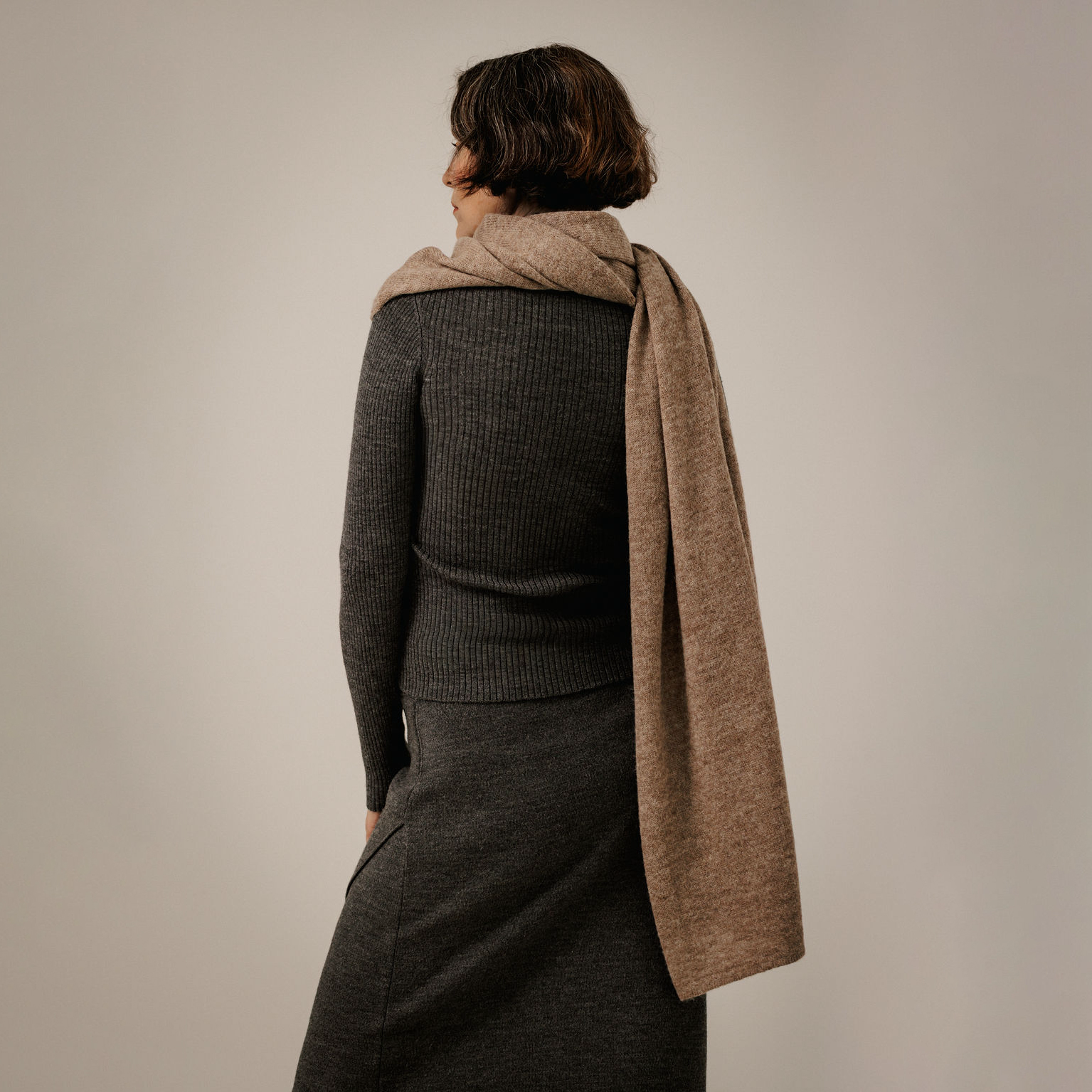 A woman shown from the back wearing a dark charcoal ribbed knit sweater and skirt, with a light brown woolen scarf draped over her shoulders, against a plain light gray studio background.