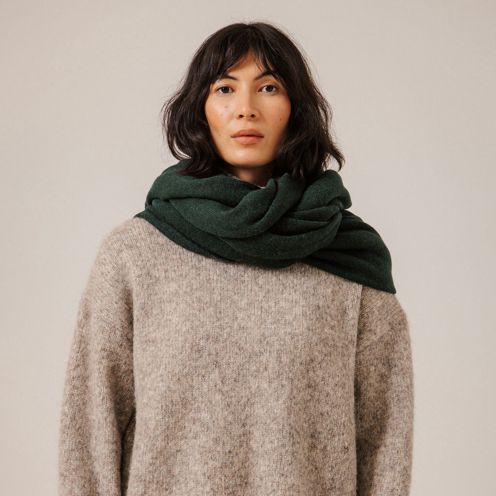 A woman with dark shoulder-length hair wearing a dark forest green knit scarf and a textured beige knit sweater, photographed against a solid light beige background.