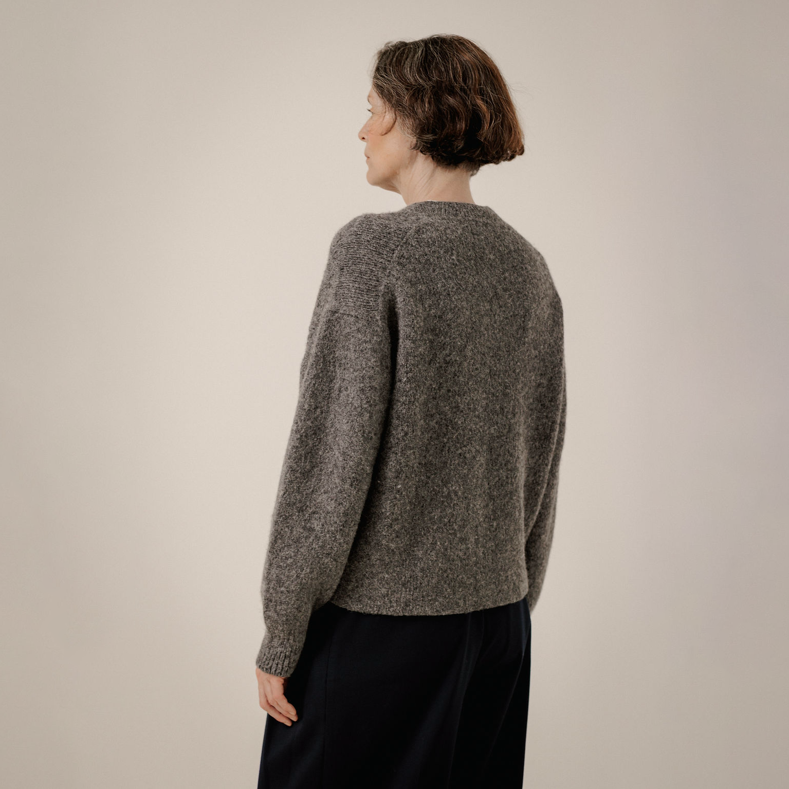 A woman shown from the back wearing a textured, heathered gray knit sweater and dark navy trousers, standing against a plain light beige studio background.