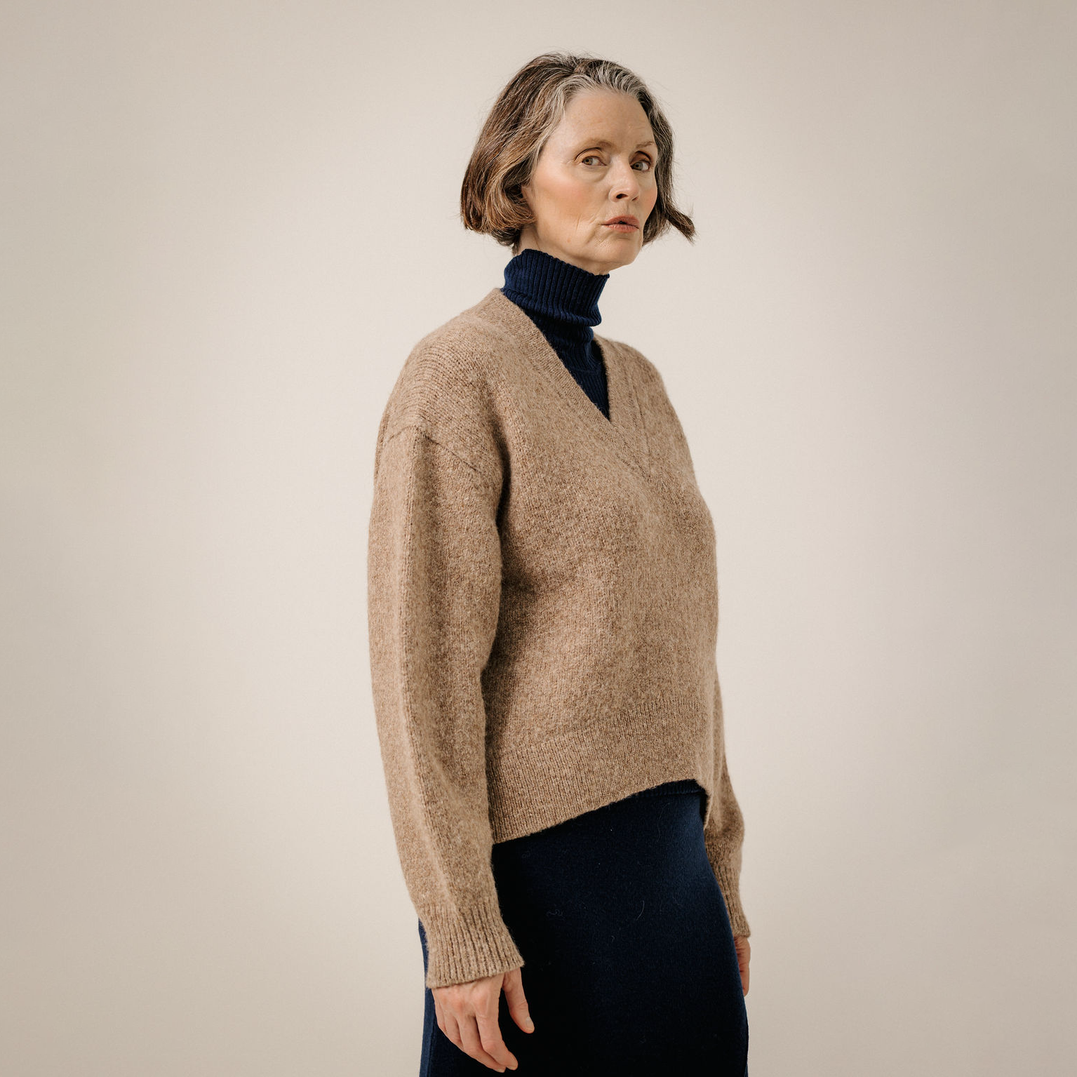 A woman wearing a beige knit V-neck sweater over a dark navy turtleneck and dark skirt, standing against a plain light beige studio background.
