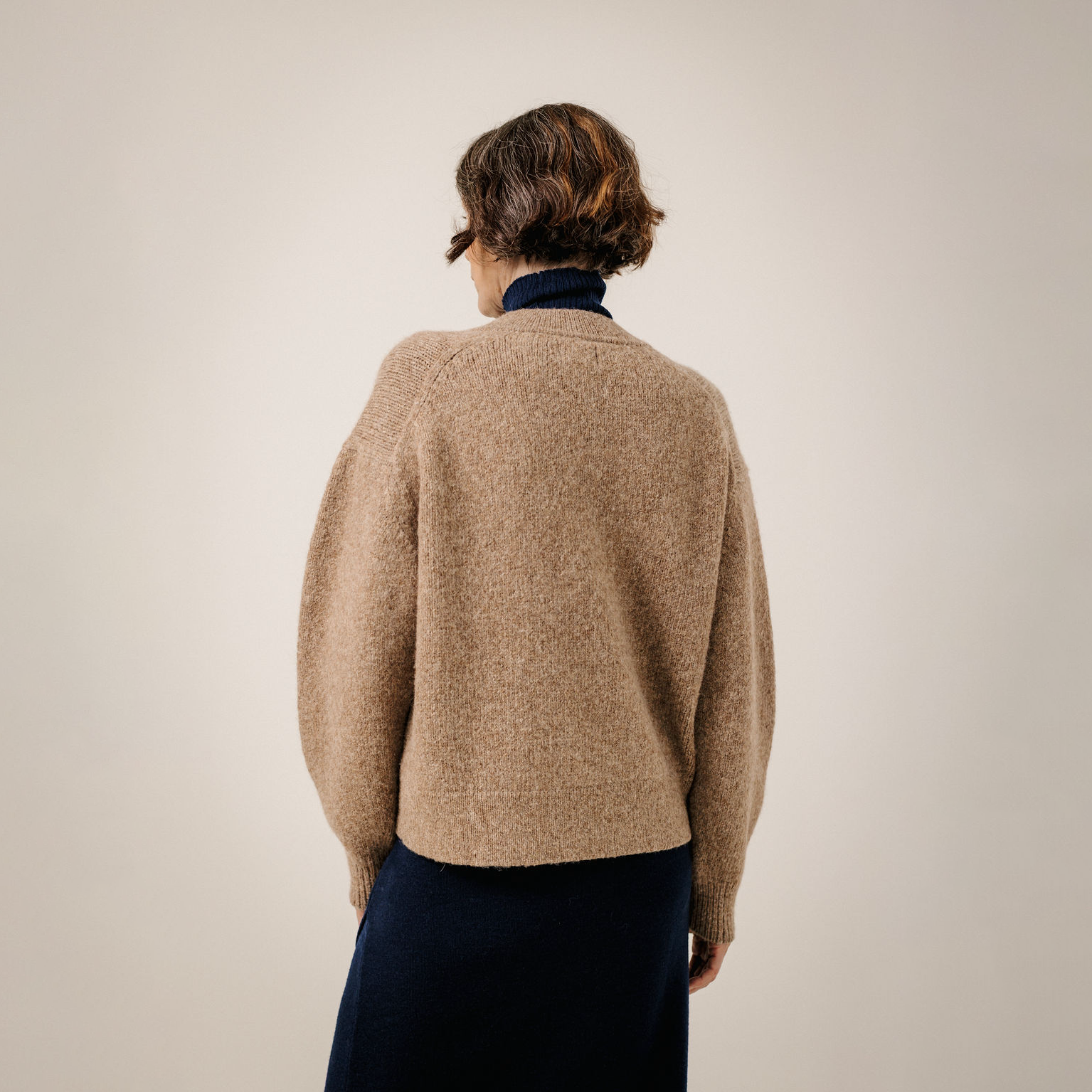 A person shown from the back wearing a textured knit sweater in a warm camel beige over a dark navy turtleneck and navy skirt, set against a plain light beige studio background.