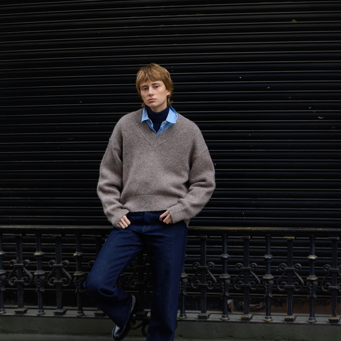 A person wearing a taupe knit sweater over a blue collared shirt and dark blue trousers, leaning against a black metal shutter background.