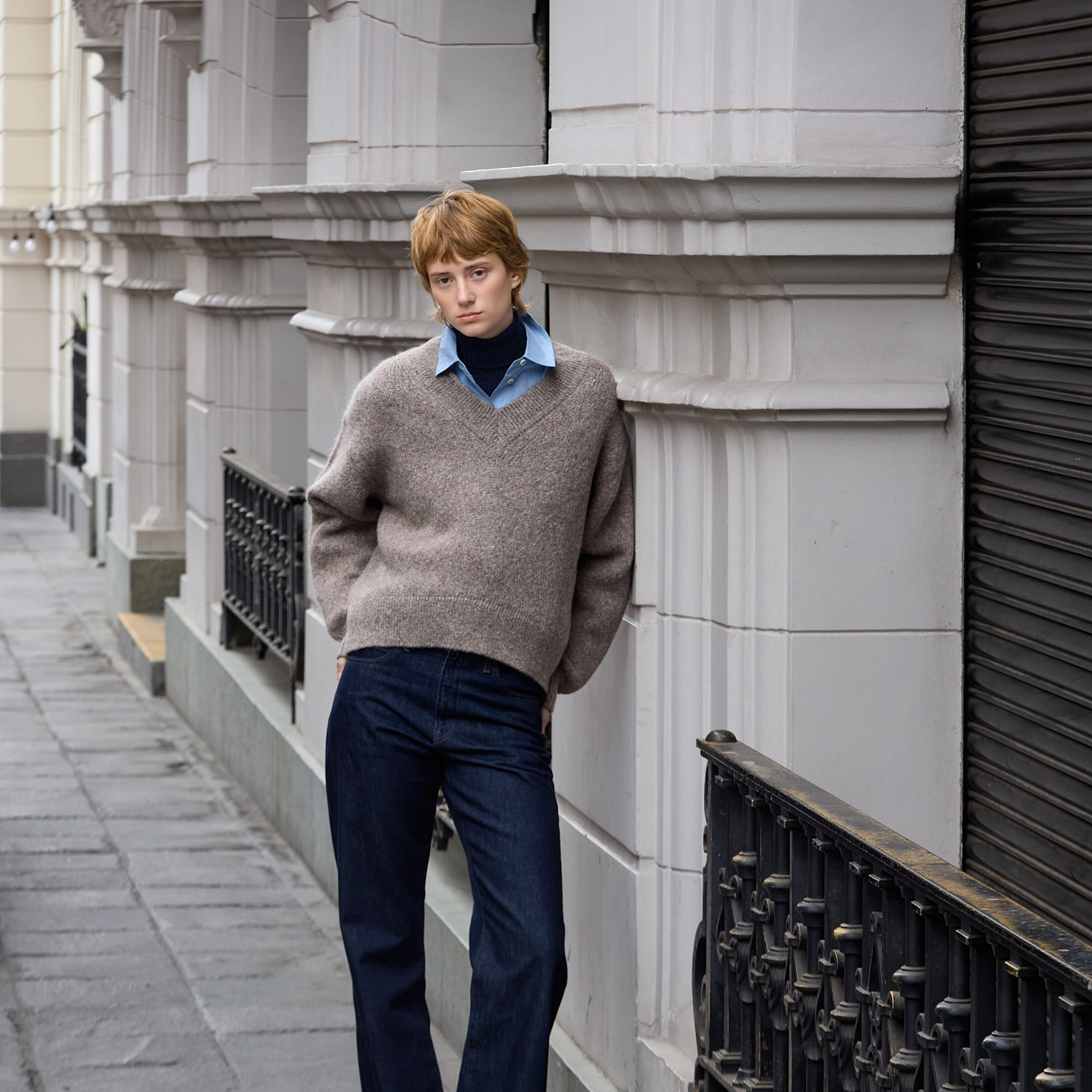 A man stands on a city sidewalk wearing a taupe knit V-neck sweater over a light blue collared shirt and dark navy trousers, posed against a white stone building facade.