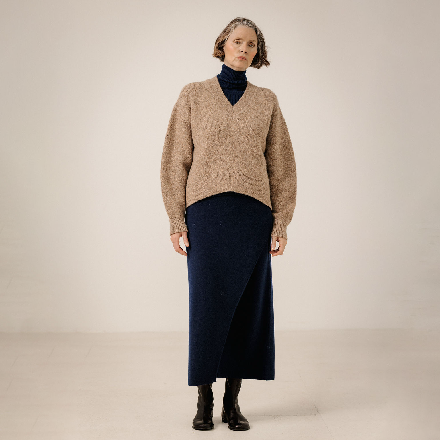 A model stands against a plain beige studio background wearing a beige knit V-neck sweater over a dark navy turtleneck, paired with a dark navy wrap skirt and black ankle boots.