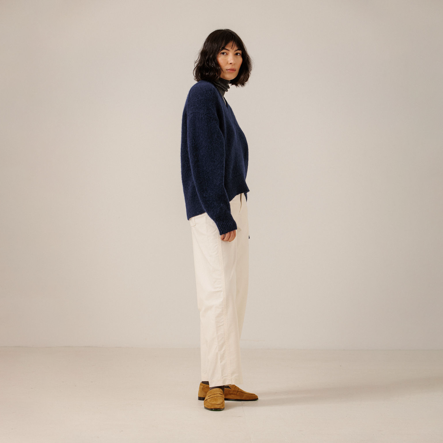 A model stands in profile wearing a navy blue knit sweater, off-white wide-leg trousers, and brown suede shoes against a plain, warm off-white studio background.