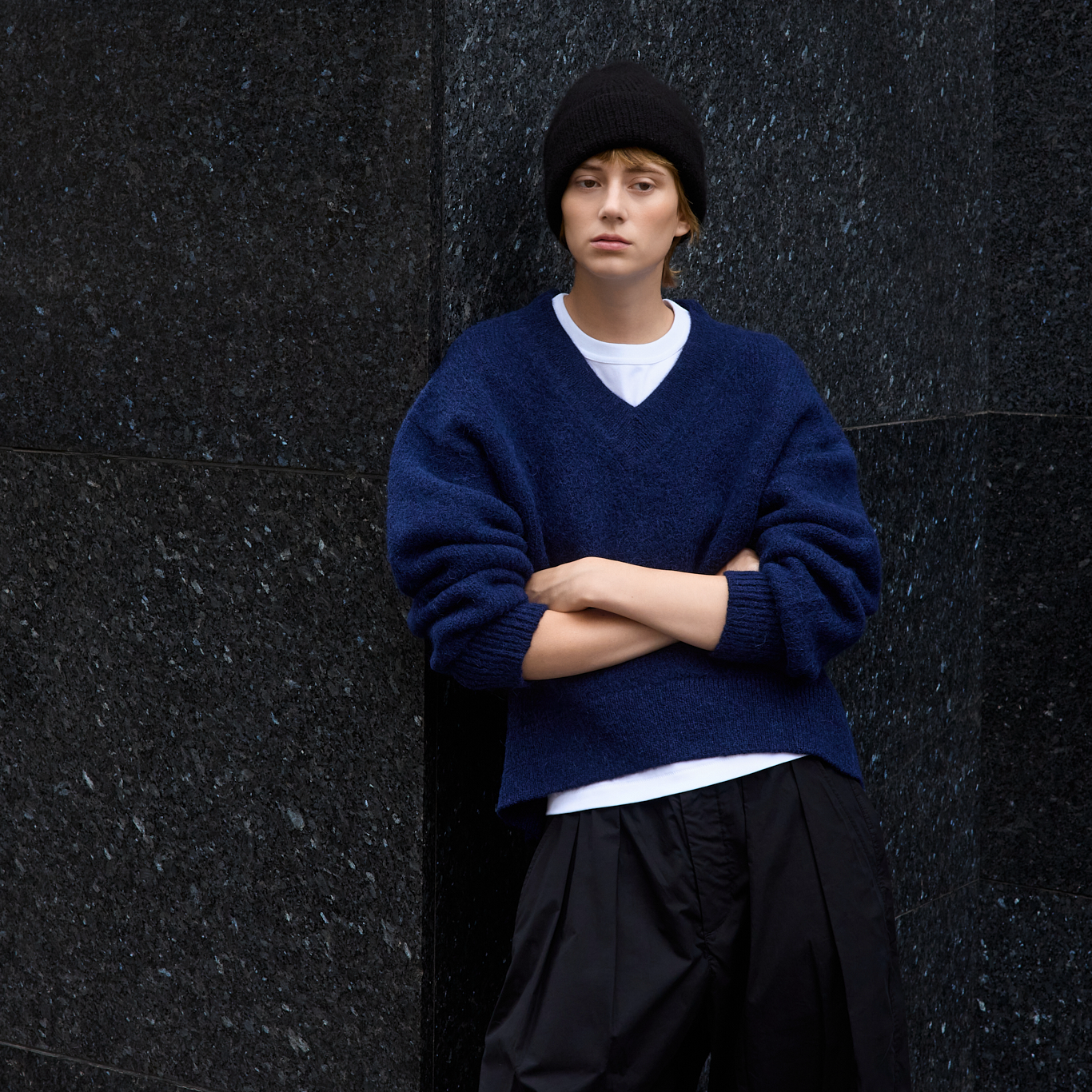 A model wearing a dark navy blue knit v-neck sweater over a white t-shirt, black pants, and a black beanie, standing against a dark textured stone wall.