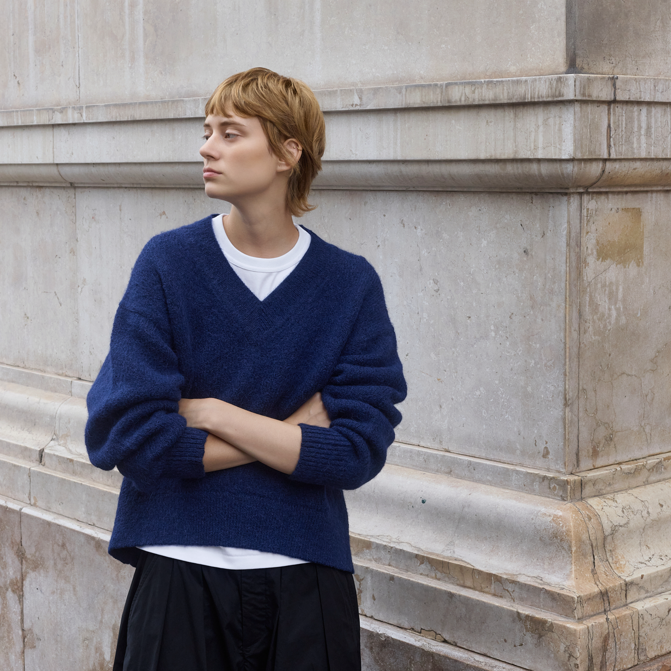 A person with short blonde hair stands against a light stone wall wearing a navy blue V-neck knit sweater over a white T-shirt and dark pants.