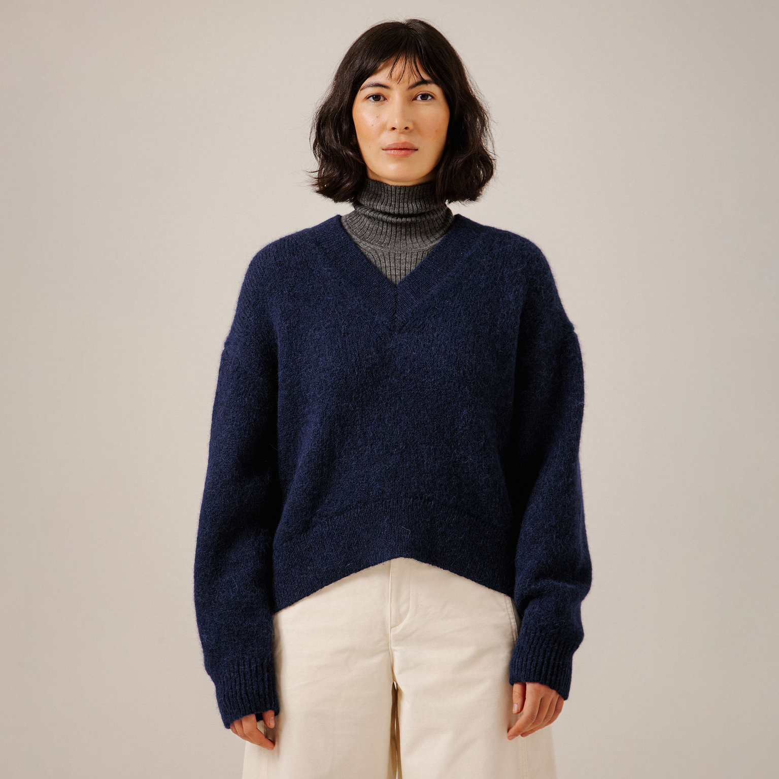 A woman wearing a navy blue V-neck knit sweater over a gray turtleneck, paired with cream pants, standing against a plain light beige studio background.
