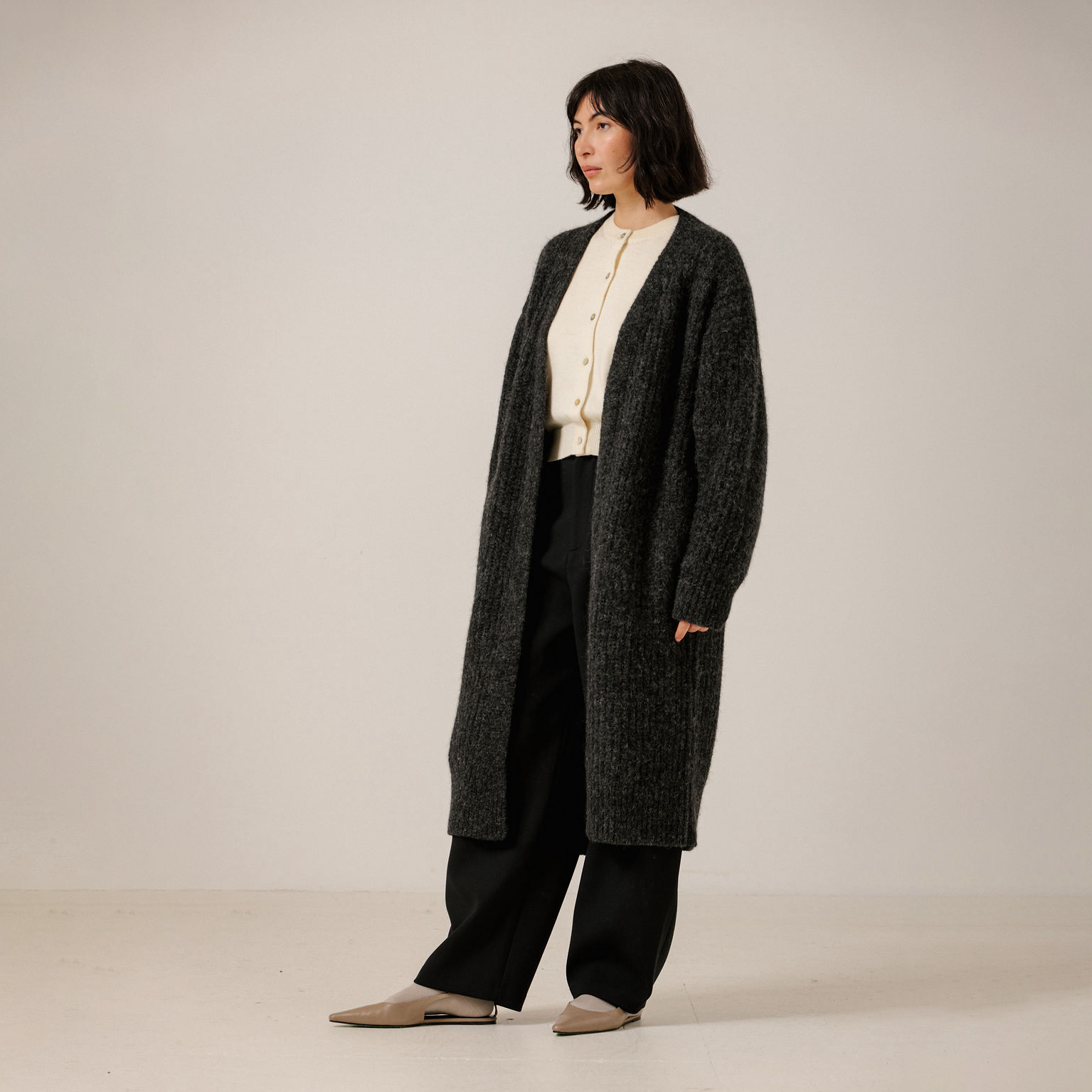 A model stands in a studio wearing a long, dark gray textured knit cardigan over a cream button-up shirt, black wide-leg pants, and beige pointed flats.