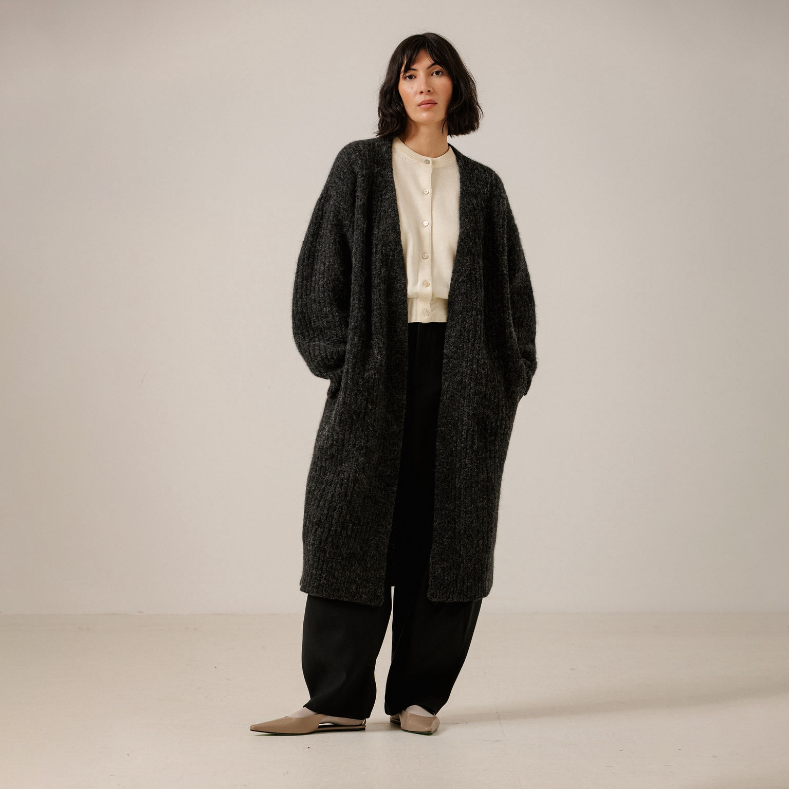 A model stands in a studio wearing a long, dark charcoal gray textured knit cardigan over a cream blouse and black wide-leg pants.