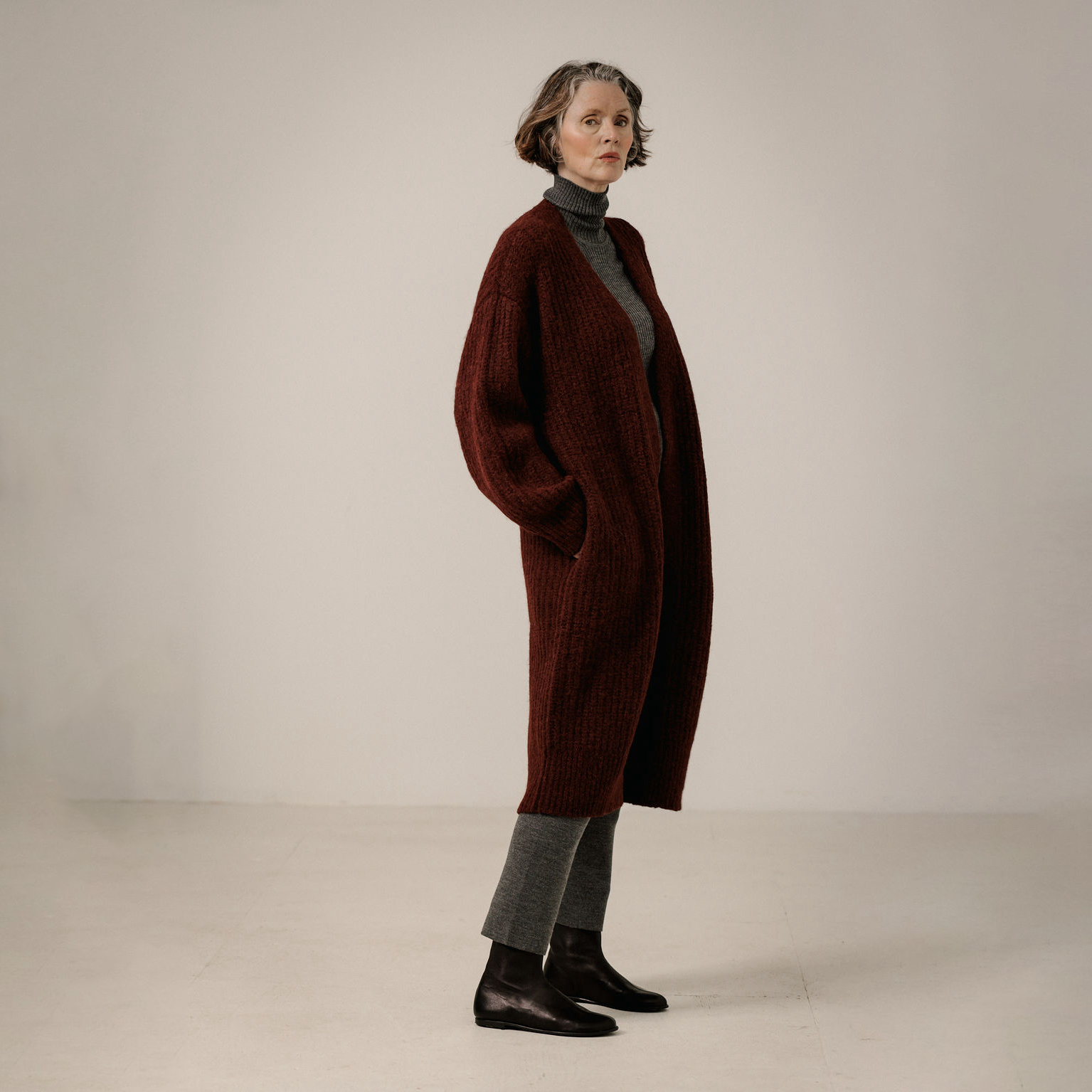 A model stands in profile wearing a long, dark burgundy ribbed knit cardigan over a dark gray turtleneck, gray knit pants, and black leather boots against a plain, light beige studio background.