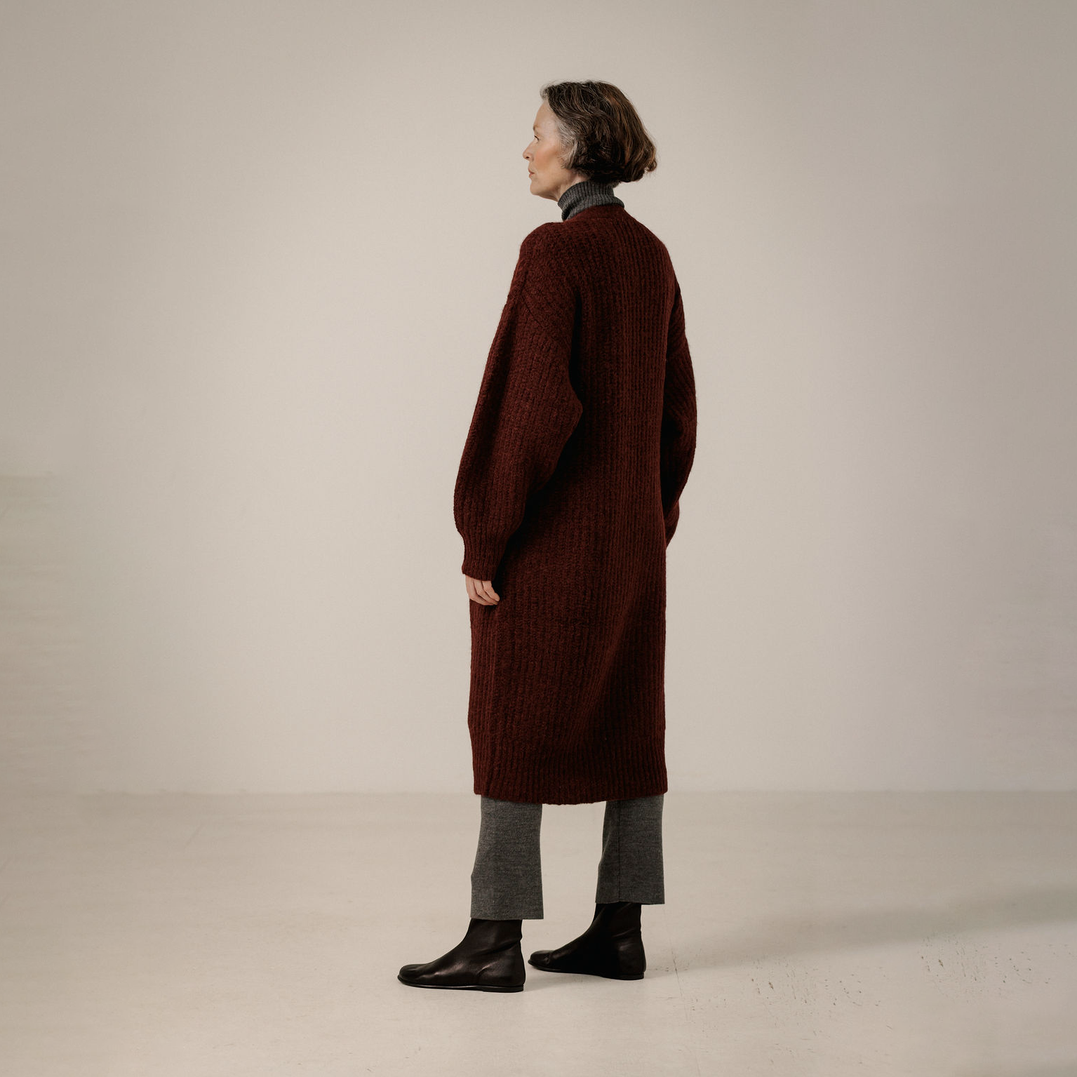 A woman stands in profile wearing a long, dark burgundy ribbed knit cardigan over a gray mock-neck top, gray trousers, and black ankle boots, photographed in a minimalist studio setting.