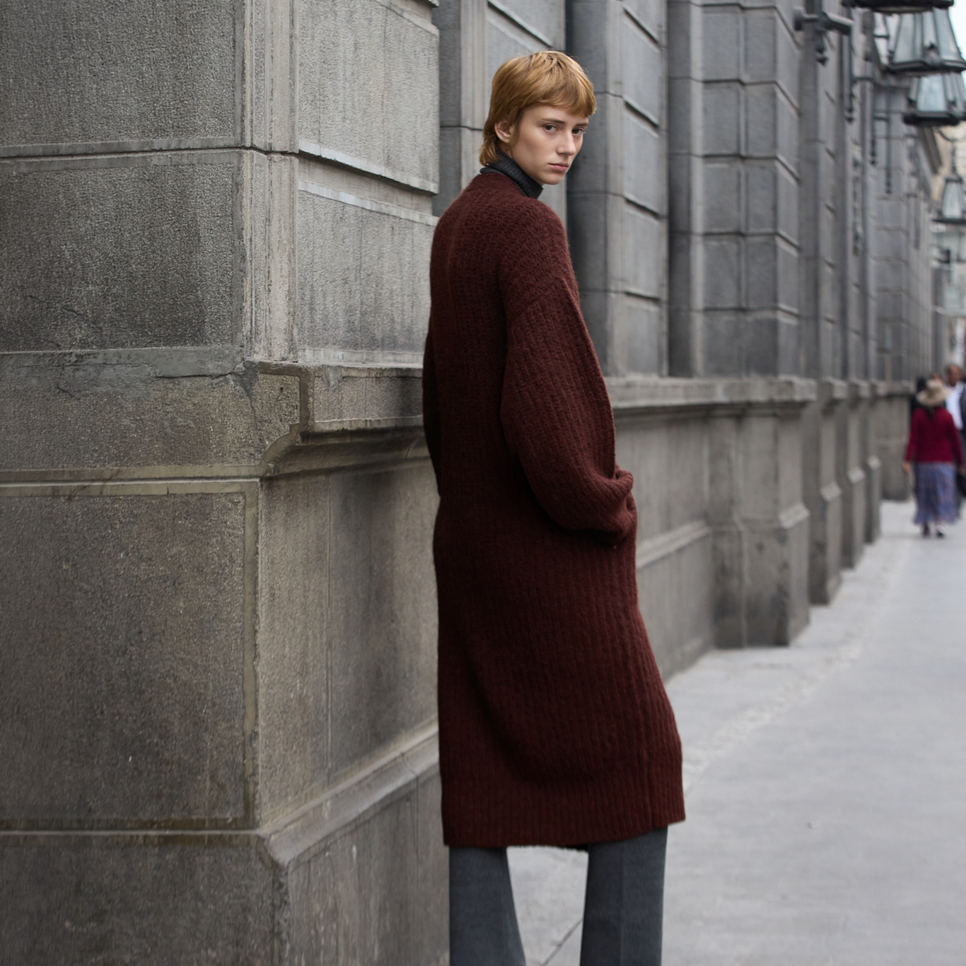 A model with red hair stands on a city sidewalk next to a gray stone building, wearing a long, dark burgundy ribbed knit cardigan over a dark turtleneck and gray pants.