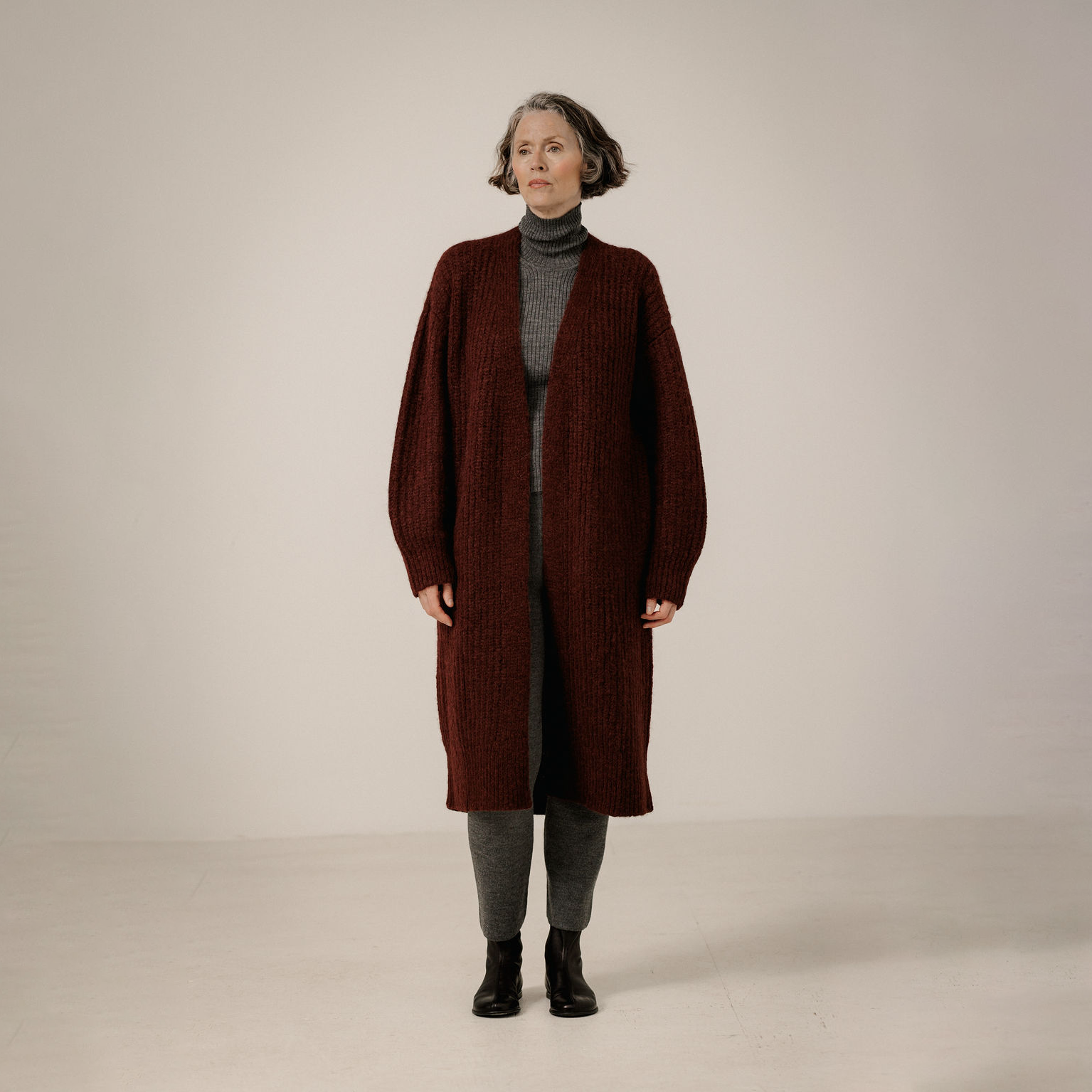 A woman stands against a plain light beige studio background wearing a long, open-front ribbed knit cardigan in deep burgundy over a gray turtleneck, gray pants, and black ankle boots.
