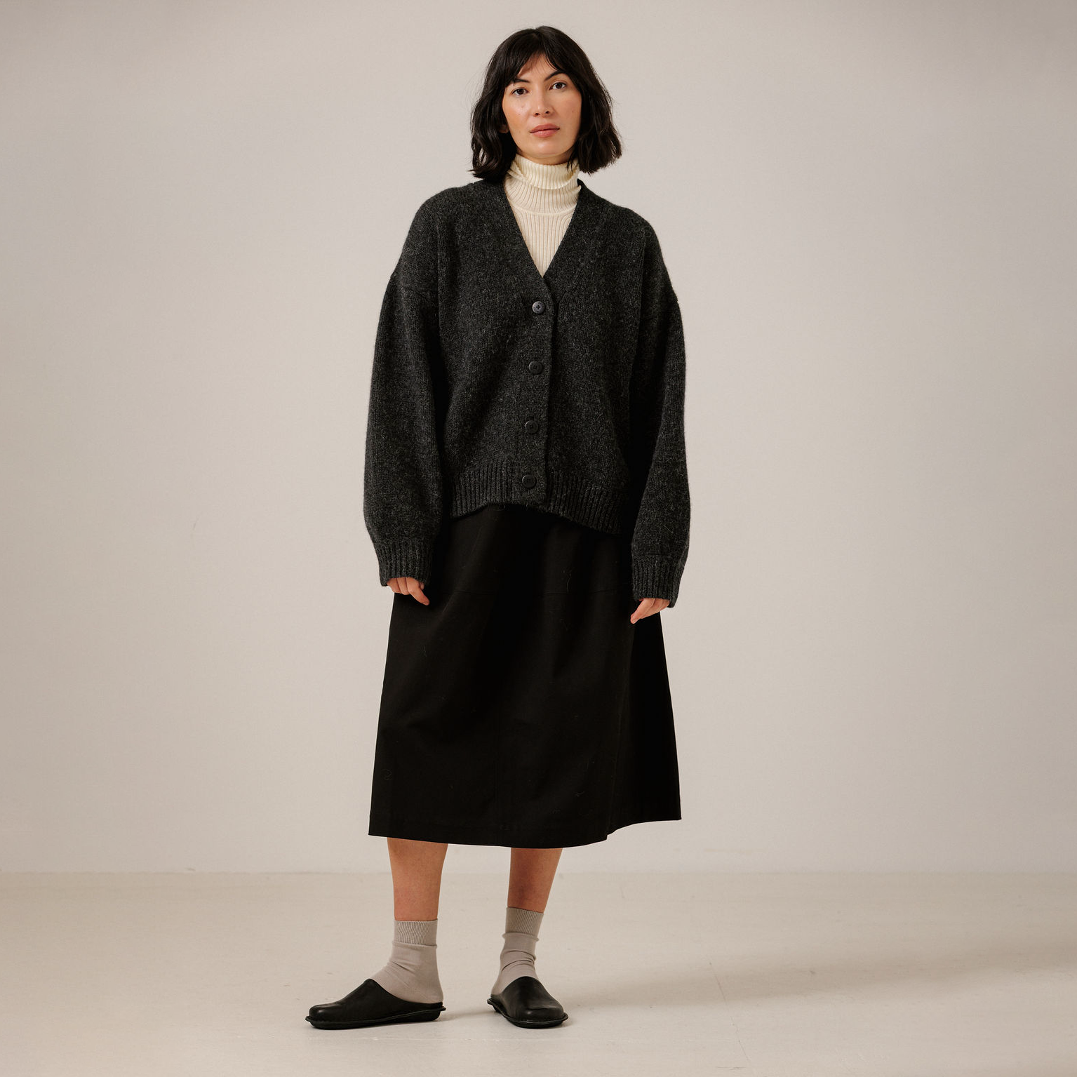 A model stands against a plain light beige studio background wearing a dark charcoal gray knit cardigan over a cream turtleneck, a long black skirt, and black slip-on shoes.