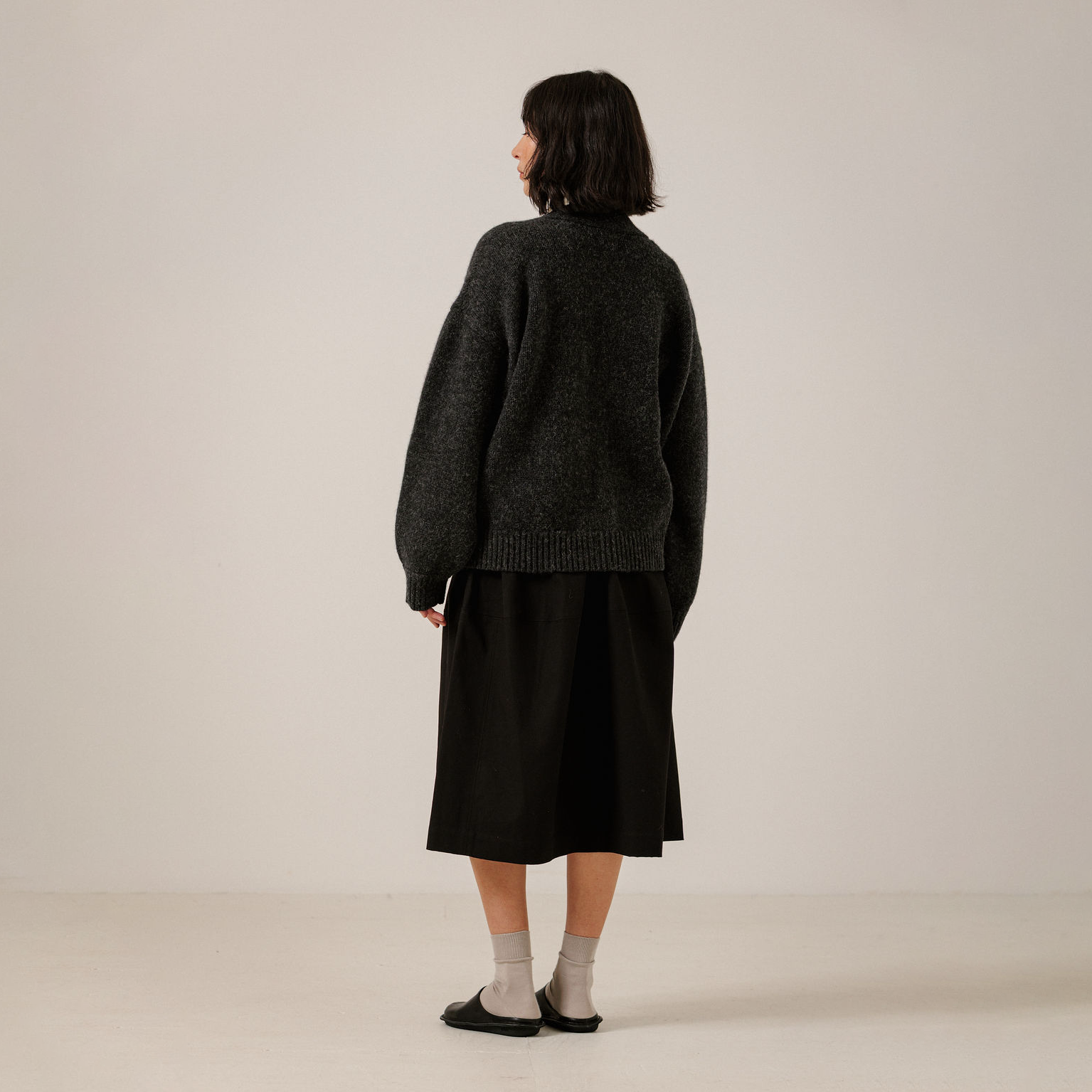 A model is shown from behind wearing a dark charcoal gray knit sweater and a black midi skirt, paired with light gray socks and black flats, against a plain off-white studio background.