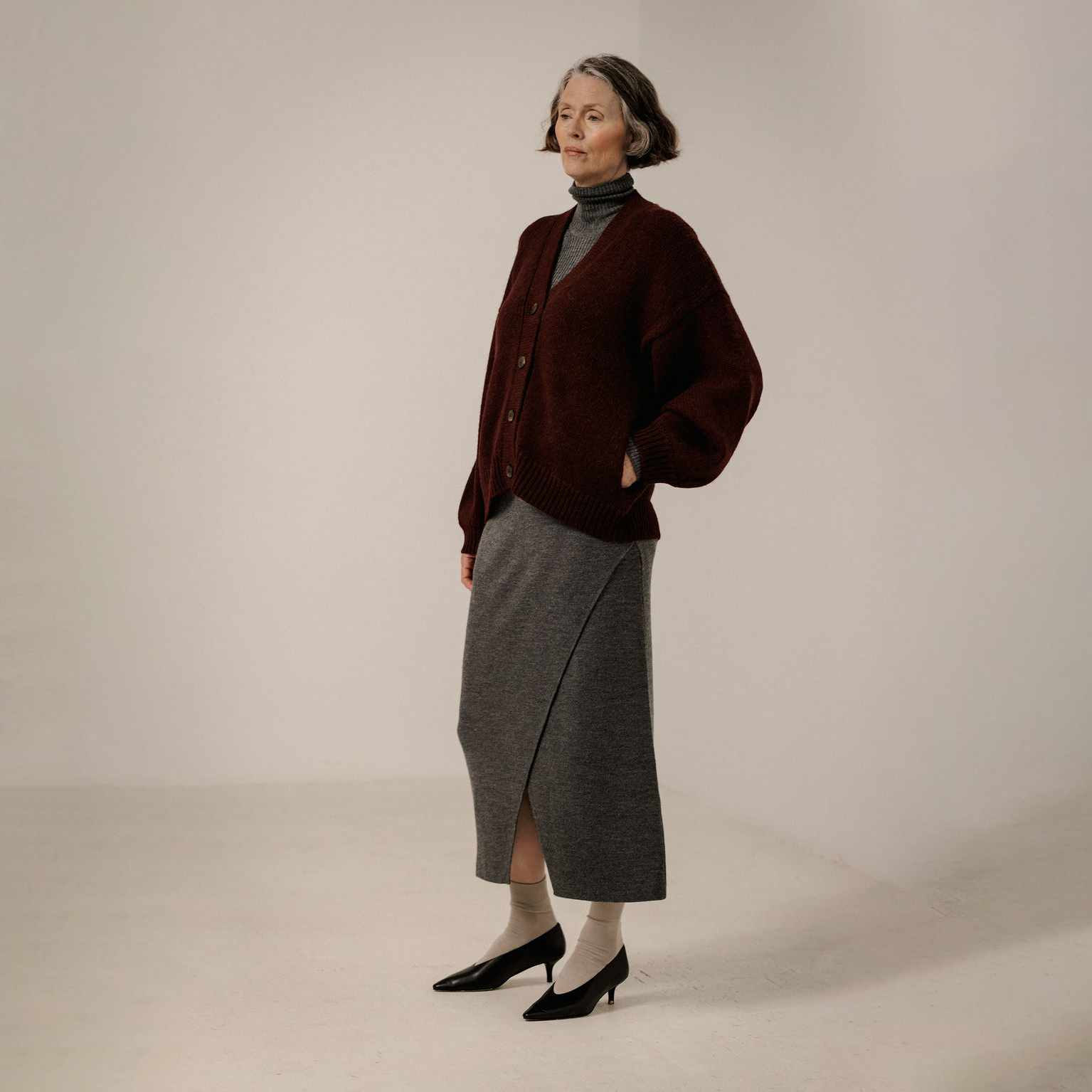 An older woman stands in a studio wearing a dark burgundy cardigan over a gray turtleneck, paired with a gray wrap skirt, white socks, and black pointed-toe heels.