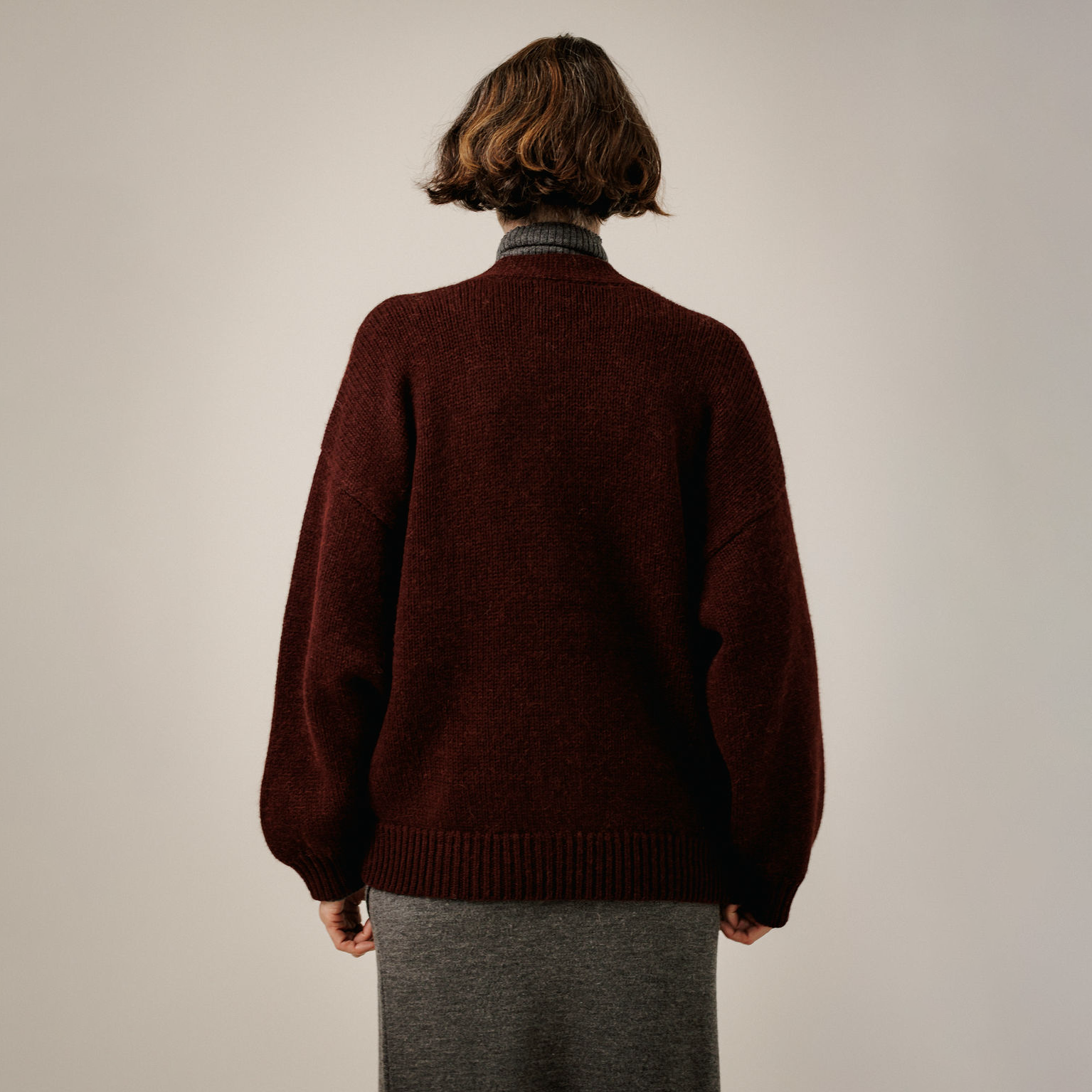 A person shown from the back wearing an oversized dark burgundy knit sweater over a gray ribbed turtleneck and gray skirt, photographed against a plain light gray studio background.