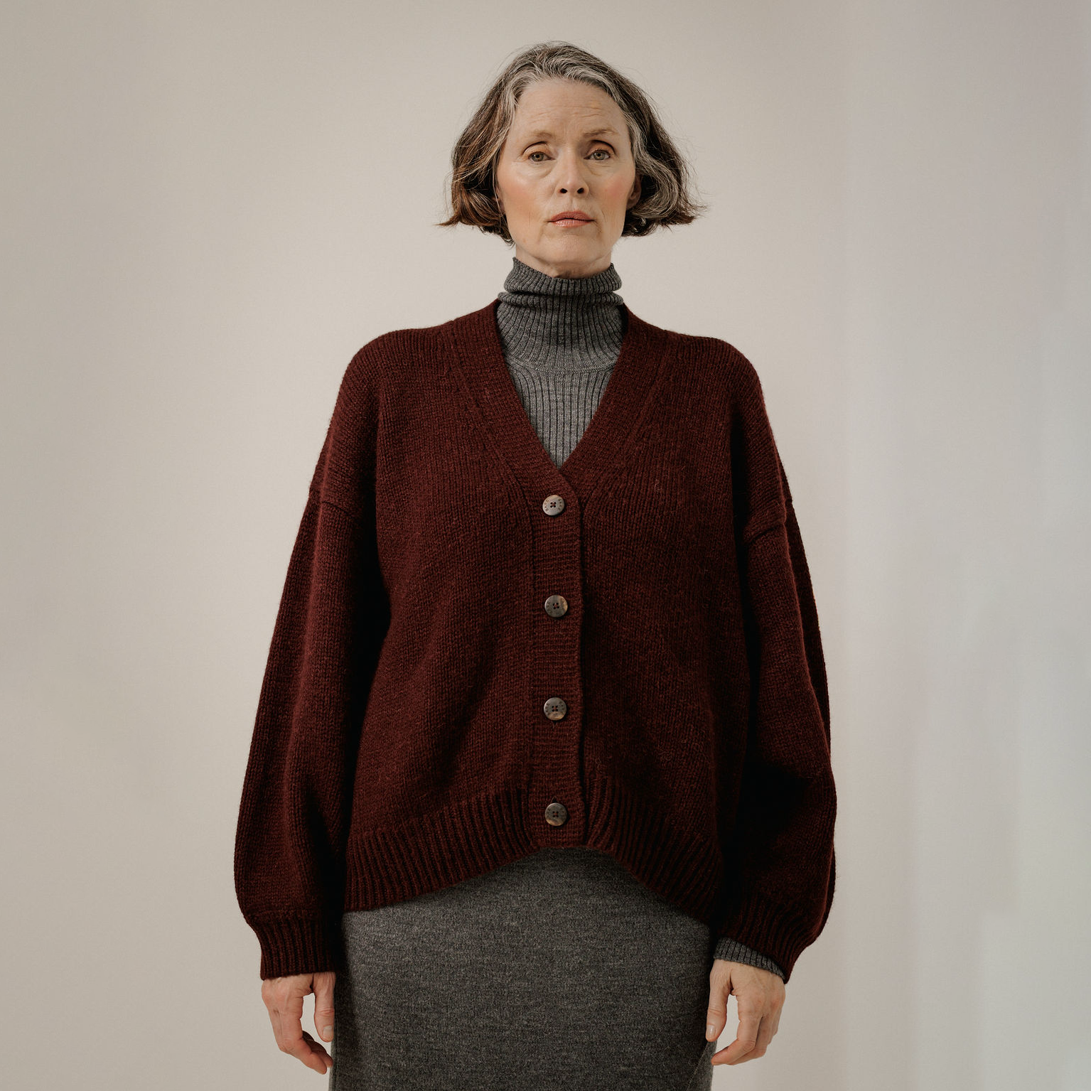 A woman wearing an oversized dark burgundy knit cardigan over a gray turtleneck and gray skirt, standing against a plain light gray studio background.