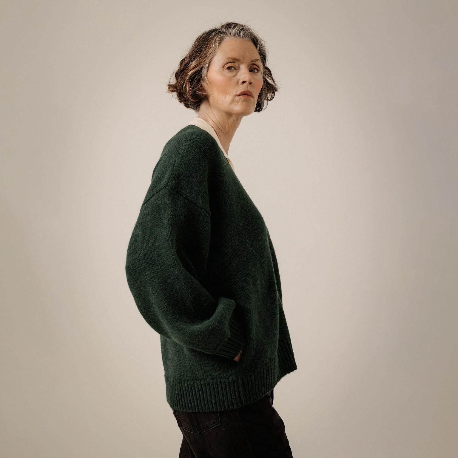 A woman wearing an oversized dark green knit sweater, shown in profile against a plain light beige studio background.