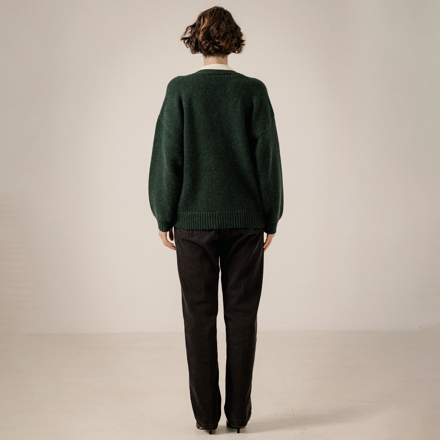 A person viewed from behind wearing an oversized dark green knit sweater and black pants, standing against a plain light beige studio background.
