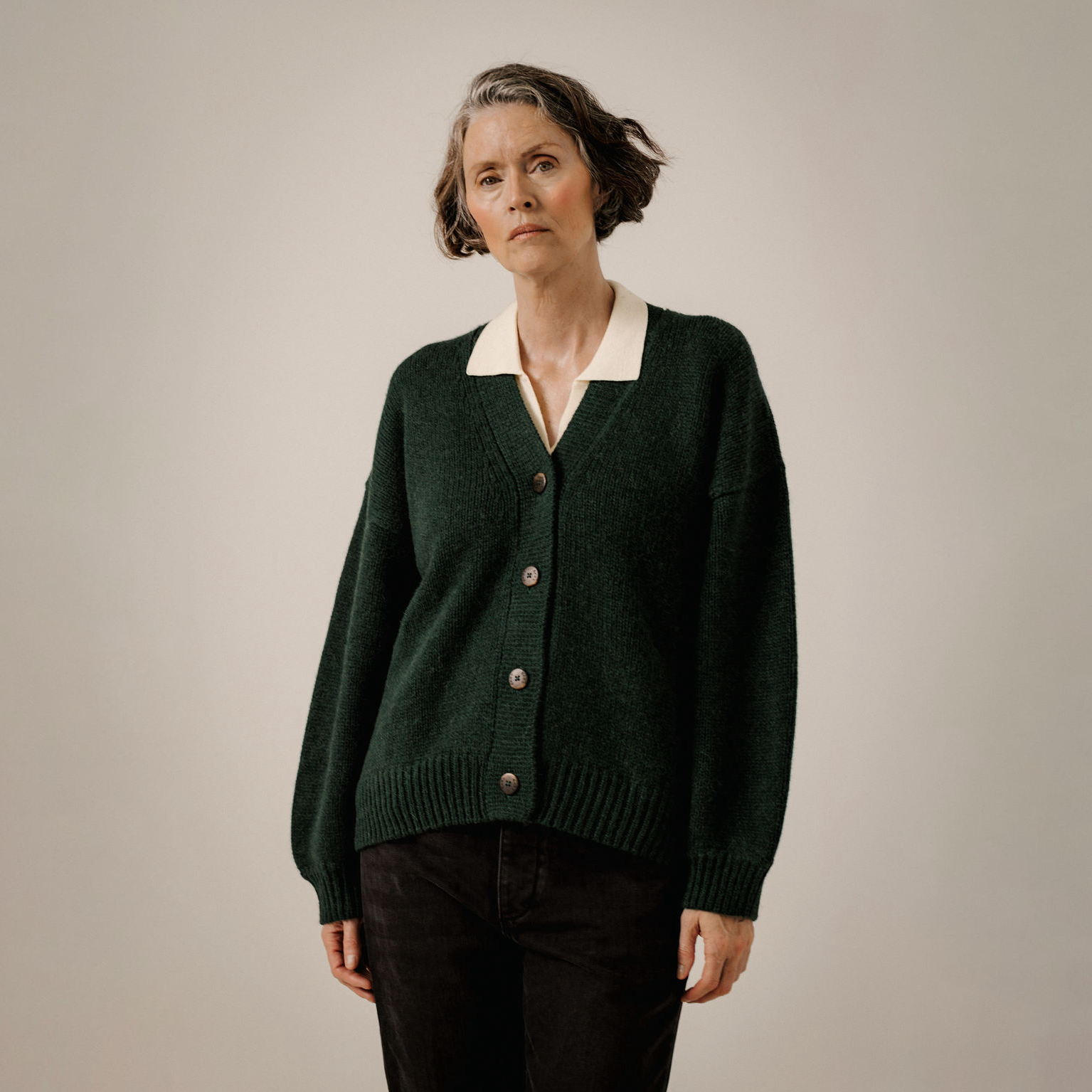 A woman wearing a dark green knit cardigan with a contrasting cream V-neck collar, paired with black pants, standing against a neutral light gray studio background.