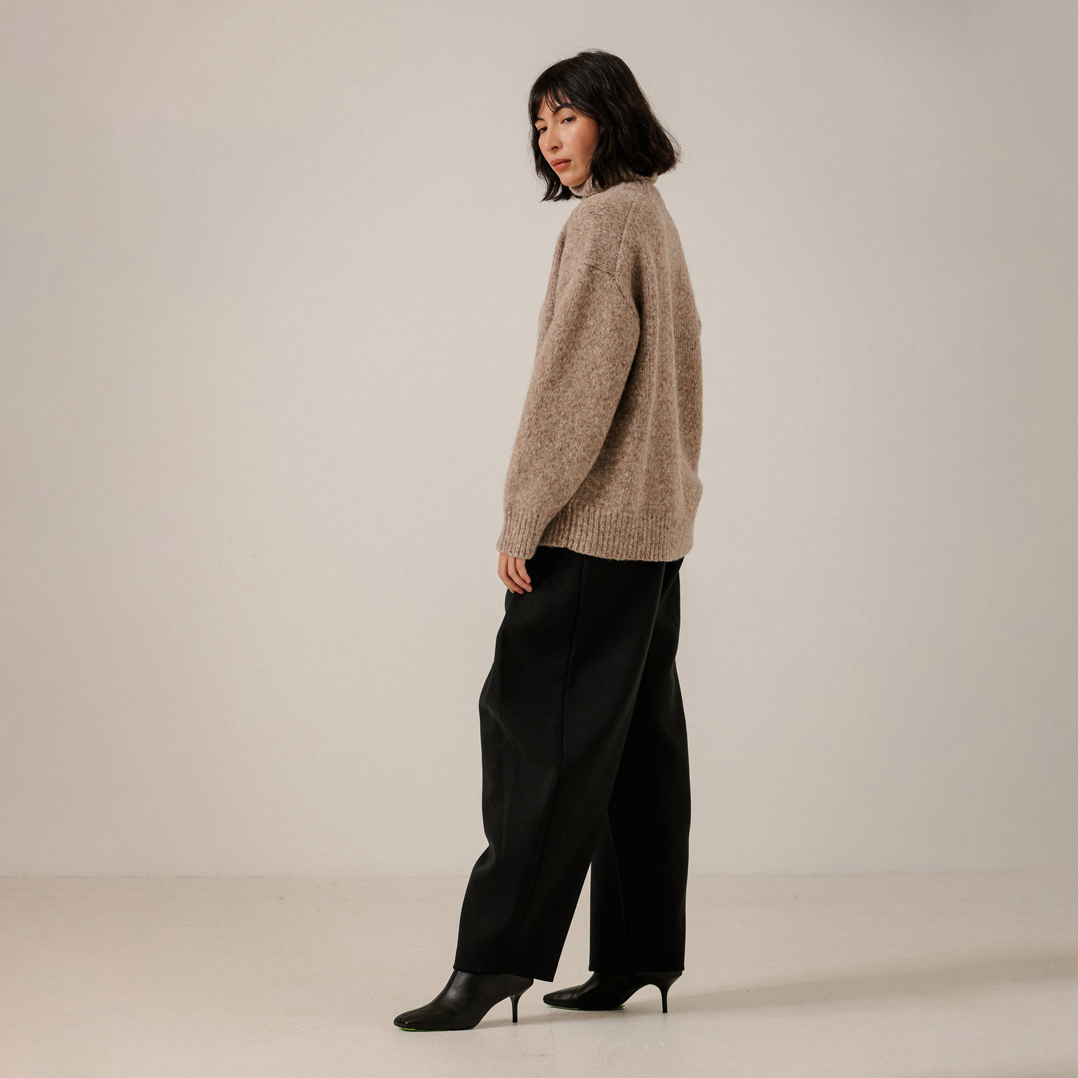 A woman stands in profile wearing a loose, fuzzy taupe knit sweater, black wide-leg trousers, and black pointed-toe heels against a plain light beige studio background.