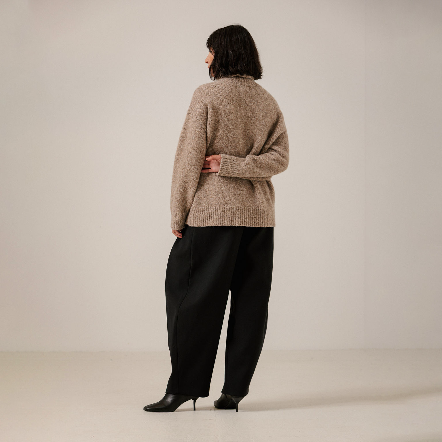 A model is shown from the back wearing a beige knit sweater and black wide-leg trousers, standing against a plain light beige studio background.
