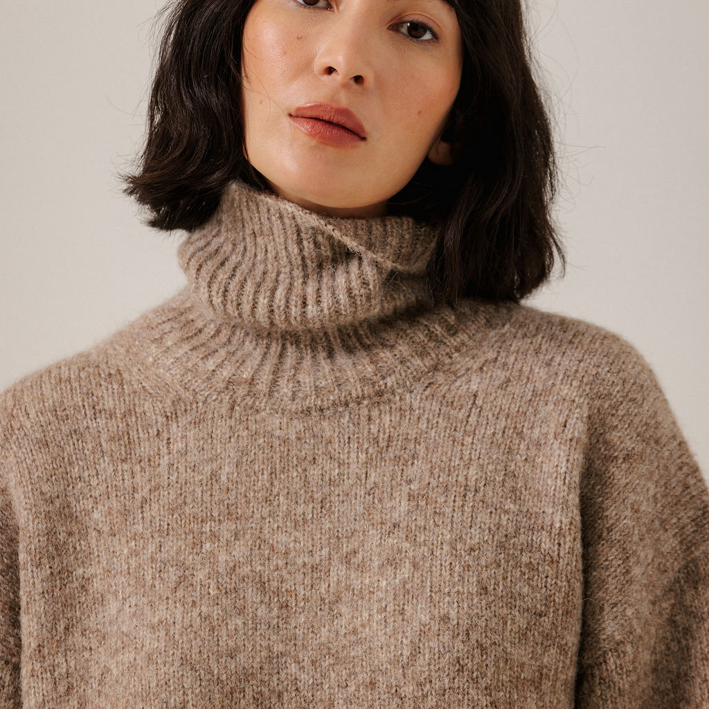 A close-up of a woman wearing a chunky, ribbed knit turtleneck sweater in a warm taupe-brown color, set against a plain light beige background.