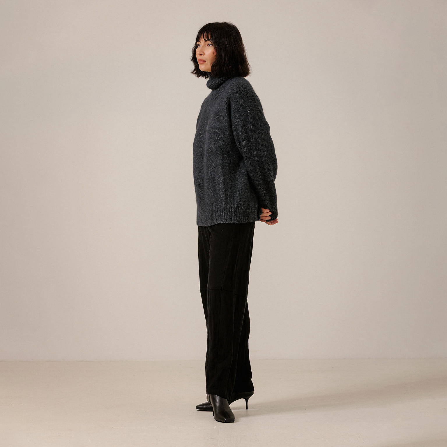 A woman stands in profile wearing a dark navy turtleneck sweater, black wide-leg trousers, and black high-heeled boots against a plain light beige studio background.