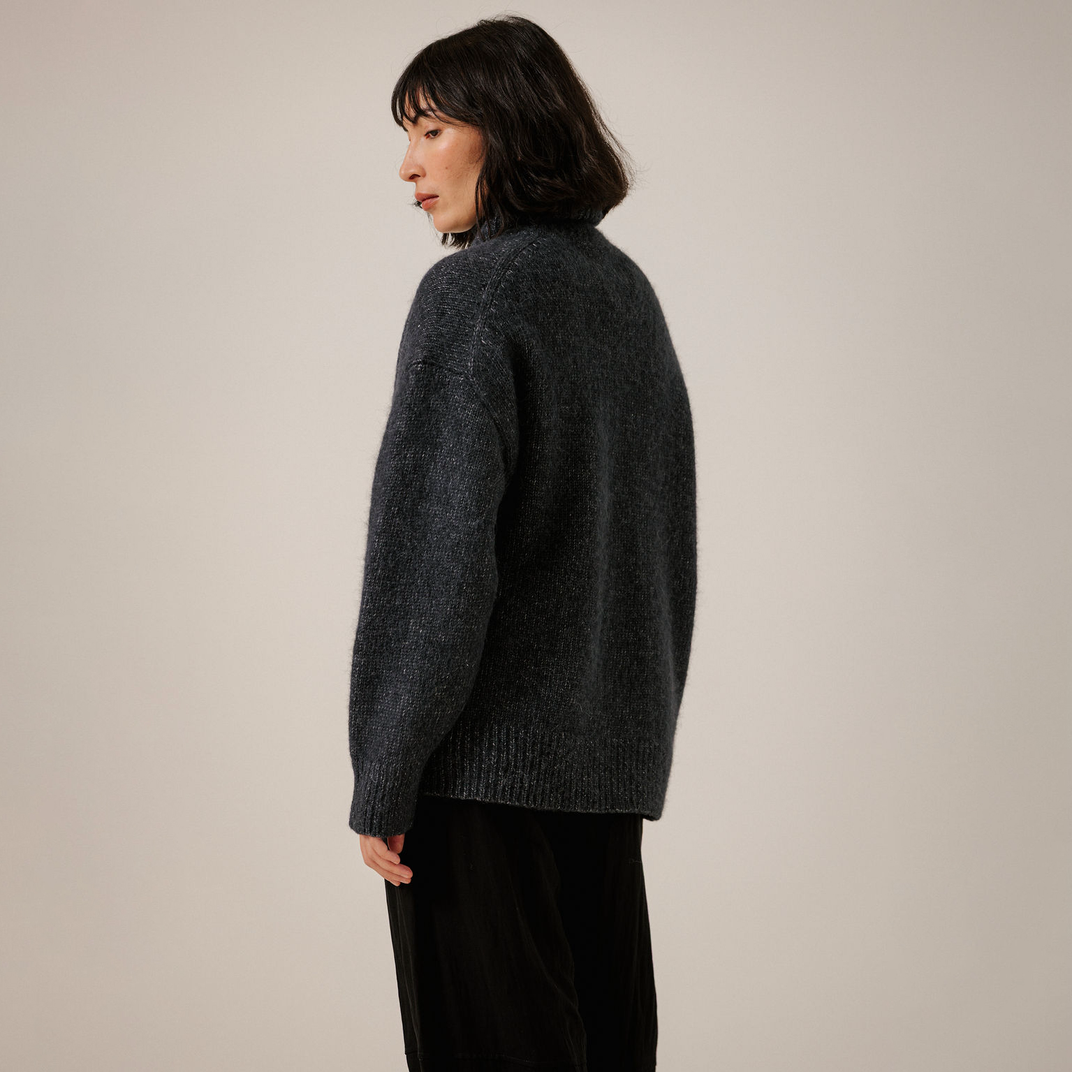 A woman shown from the back wearing a dark navy fuzzy knit sweater and black pants, standing against a plain light beige studio background.