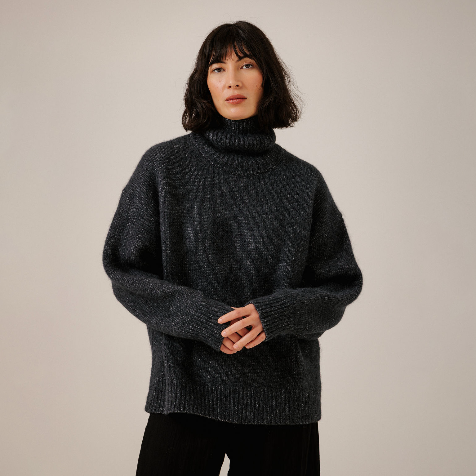 A woman wearing a dark navy knit turtleneck sweater with a chunky ribbed collar, cuffs, and hem, paired with black pants, standing against a plain light beige studio background.