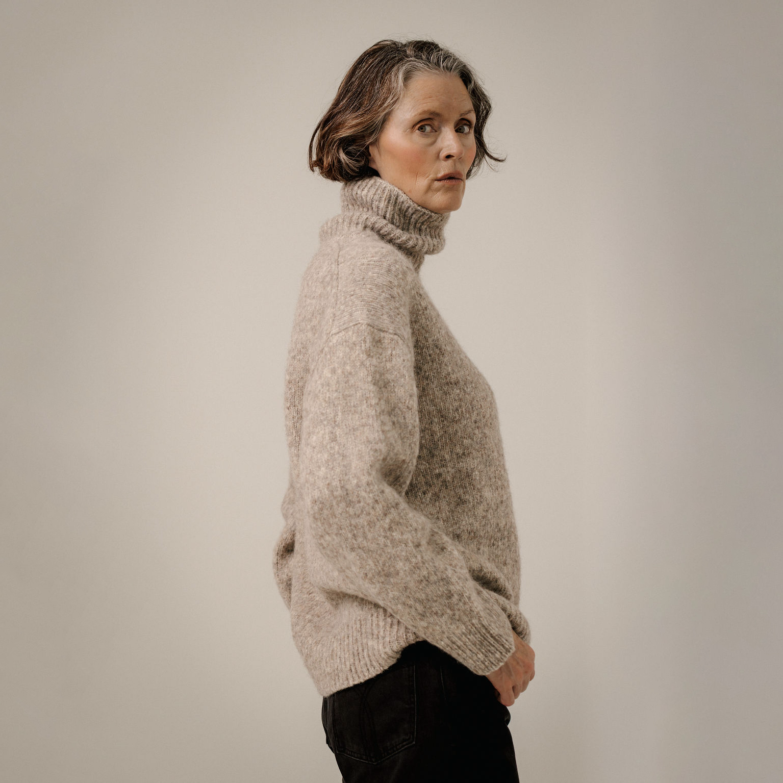 A woman wearing a chunky, textured knit turtleneck sweater in a heathered beige color, paired with black pants, photographed against a neutral light gray studio background.