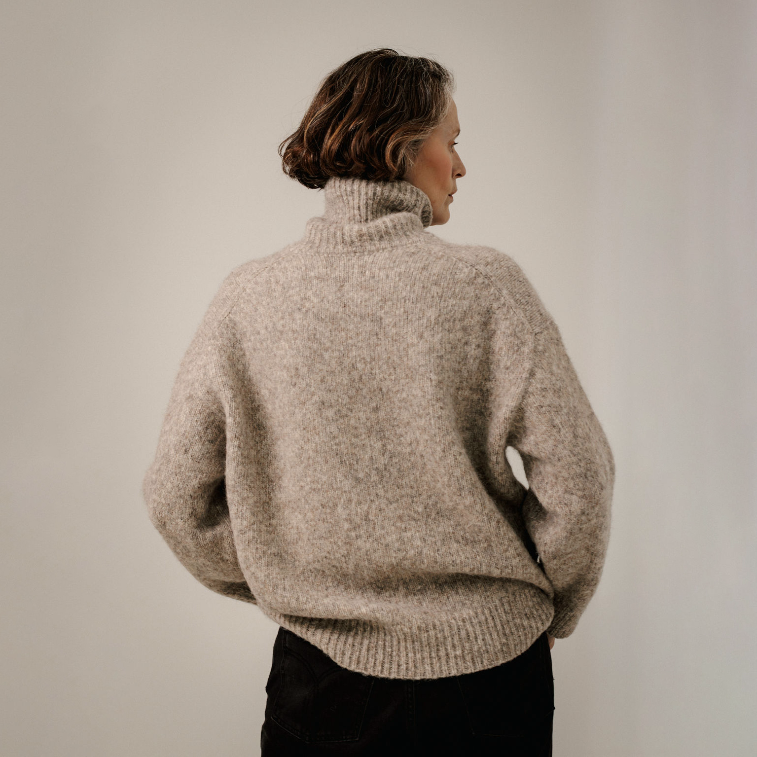 A woman shown from the back wearing a chunky, textured turtleneck sweater in a heathered light brown/beige color, paired with black pants, against a plain light gray studio background.