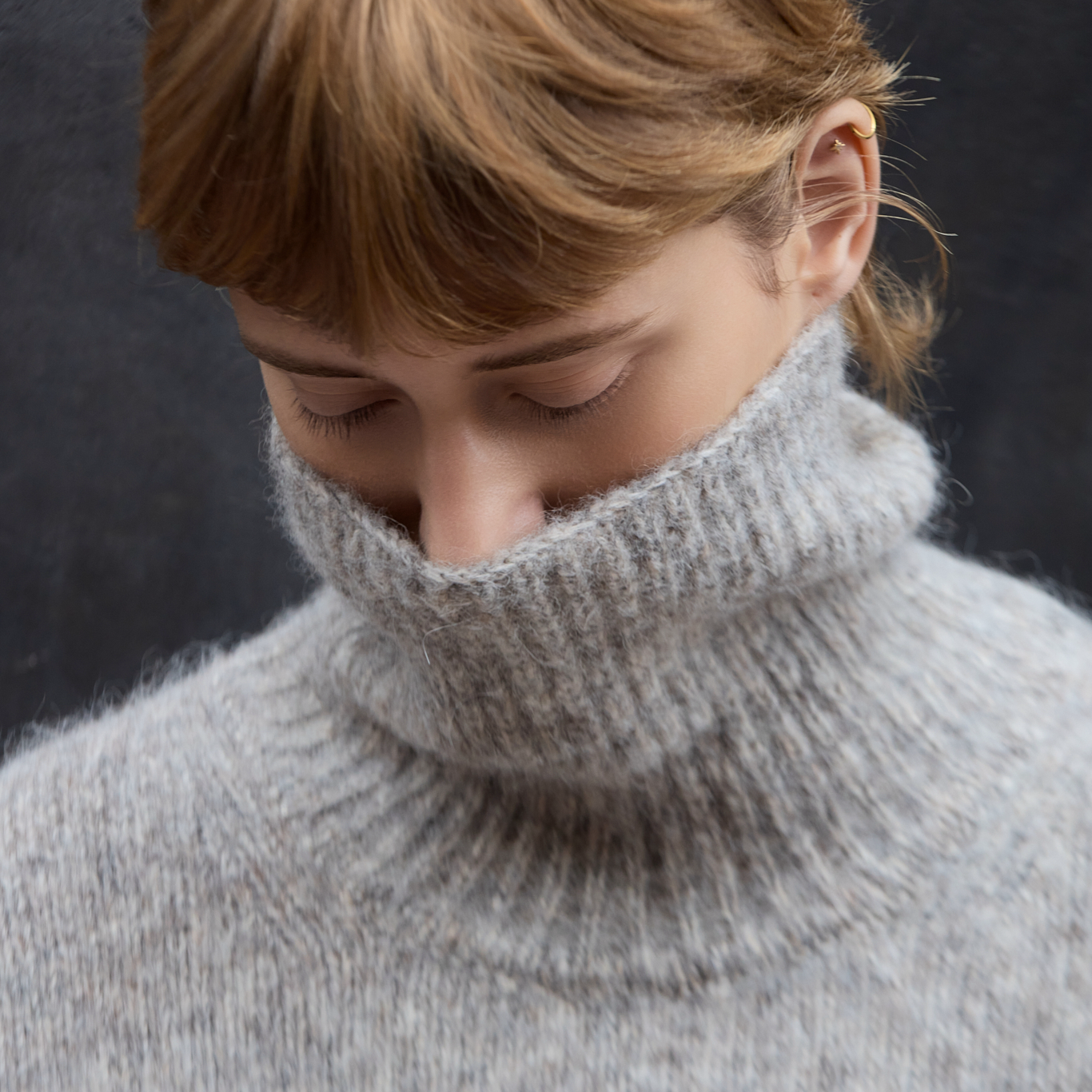 Close-up of a person wearing a soft, fuzzy light gray turtleneck sweater, looking down against a dark gray background.