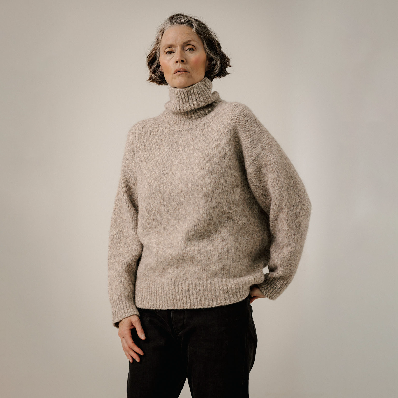 An older woman wearing a chunky, oversized turtleneck sweater in a heathered oatmeal-beige color, paired with black pants, standing against a neutral light gray studio background.