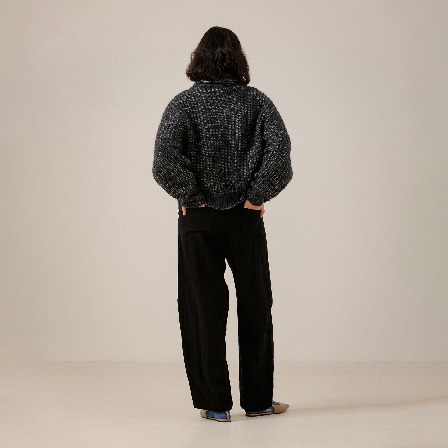 A person shown from the back wearing a dark navy ribbed knit sweater and black wide-leg trousers, standing against a plain off-white studio background.