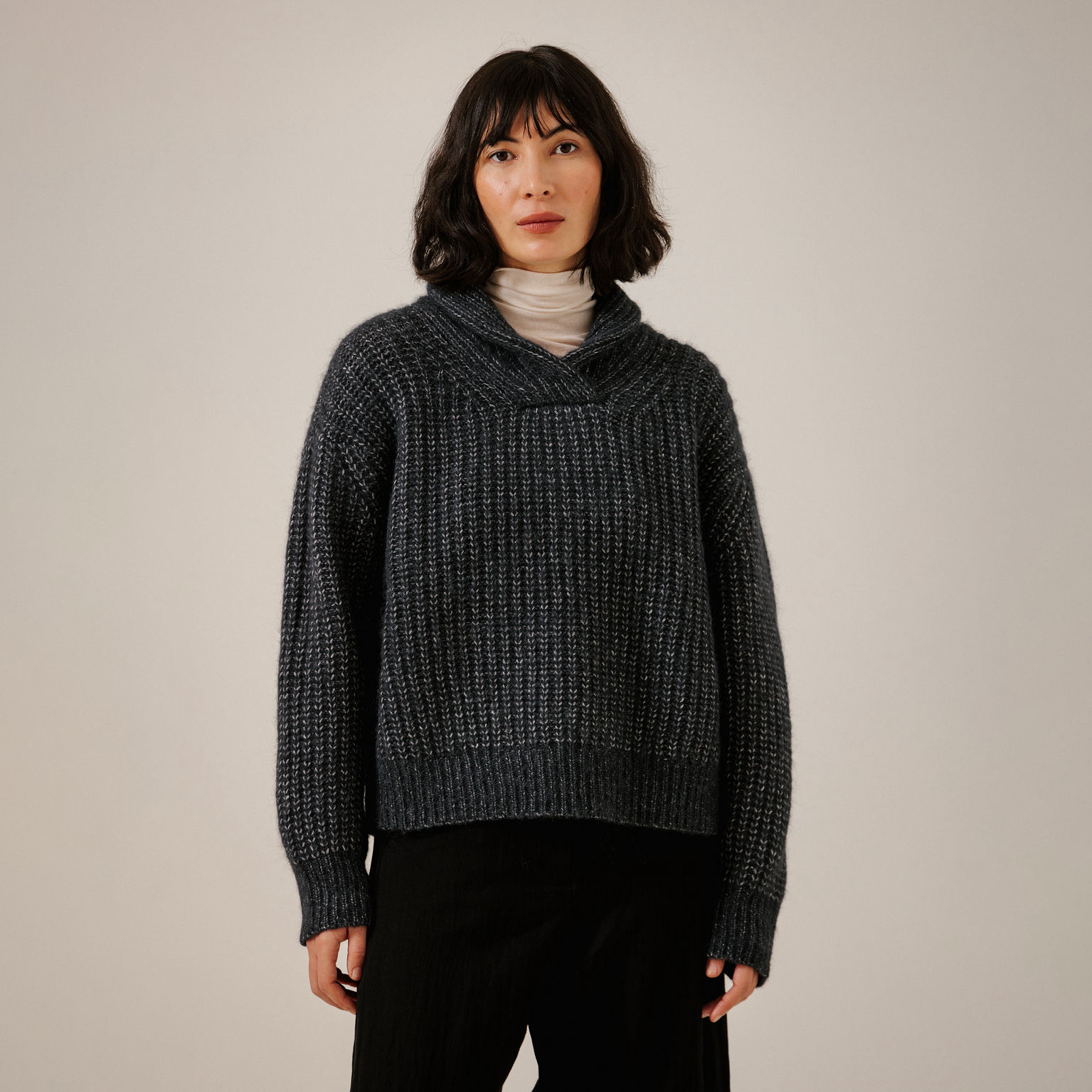 A woman wearing a dark navy textured knit sweater with a wide cowl neck, layered over a light beige turtleneck and black pants, standing against a plain light gray studio background.