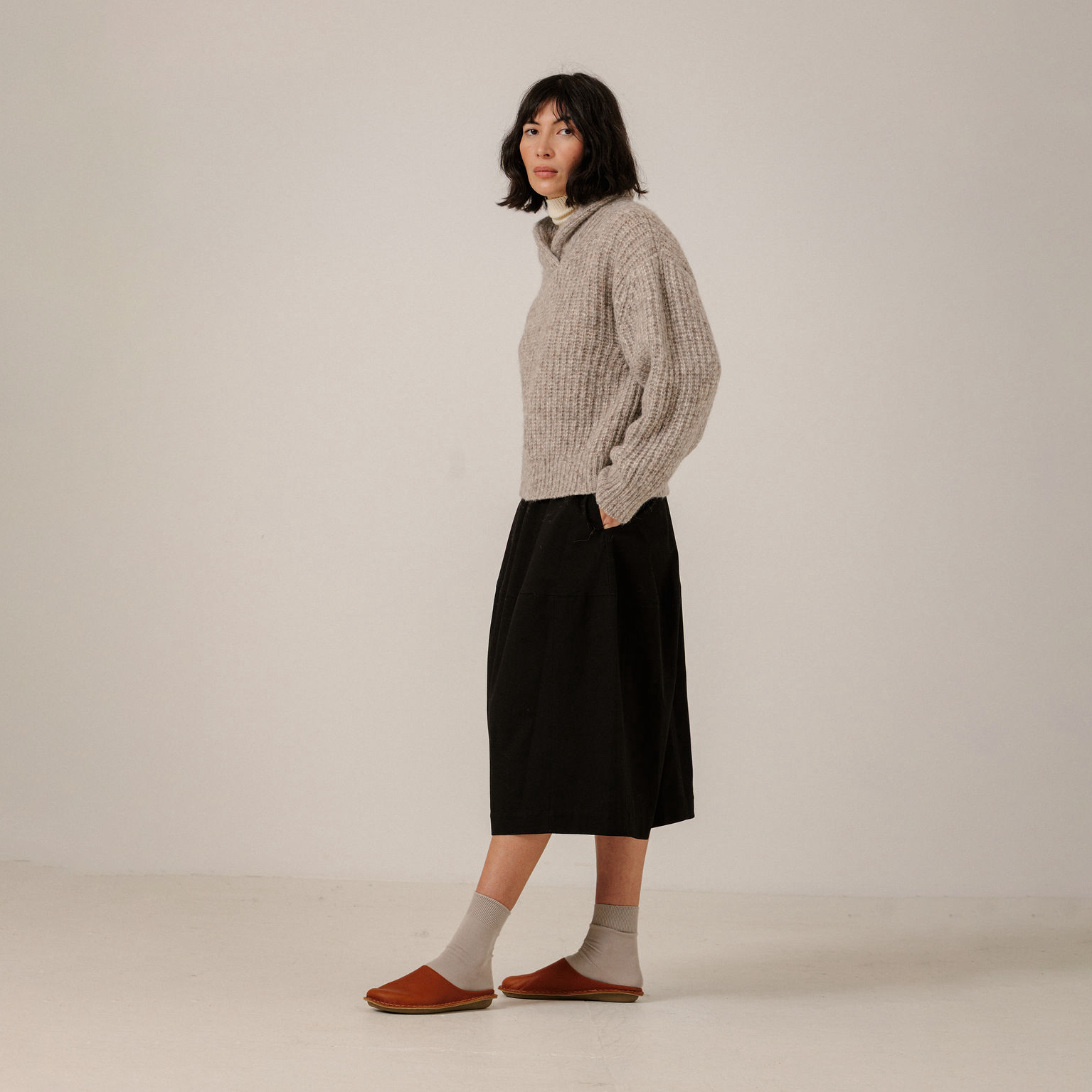 A model stands in profile wearing a chunky beige/grey shawl collar sweater, a black midi skirt, beige socks, and brown slip-on shoes against a plain off-white studio background.