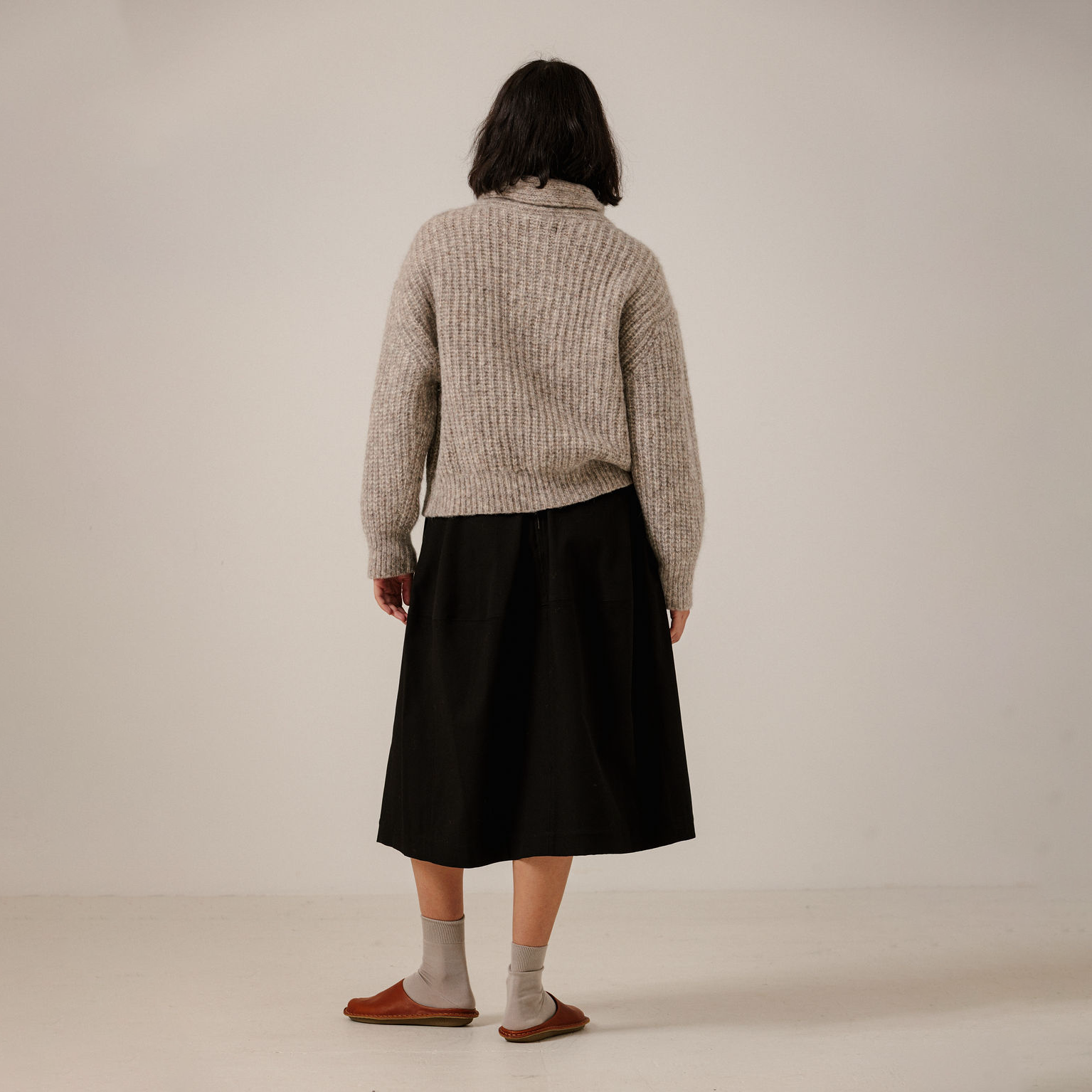 A person standing with their back to the camera, wearing a beige ribbed knit sweater, a black midi skirt, gray socks, and brown slip-on shoes against a plain light beige studio background.